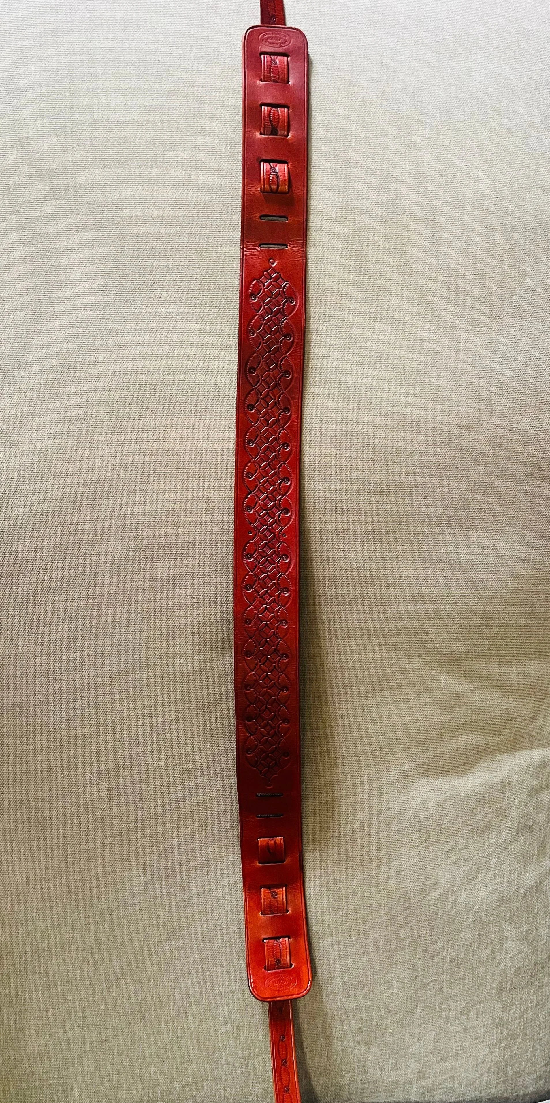 Red leather belt with ornate carved design, hanging vertically on a beige fabric background.