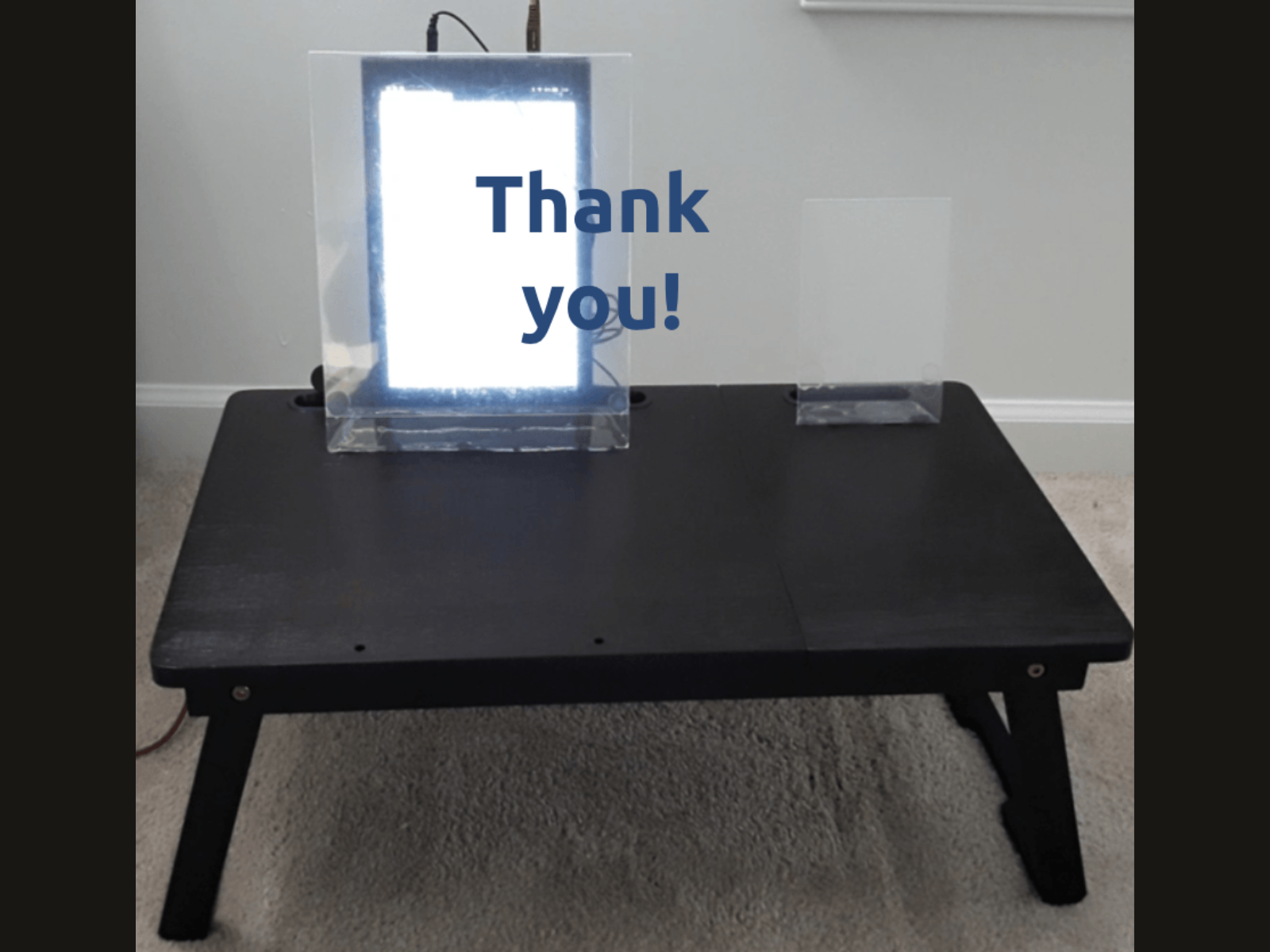 A black table with a clear acrylic sign that says "Thank you!" in blue letters, positioned in front of a bright LED light and a small transparent acrylic stand on a beige carpeted floor.