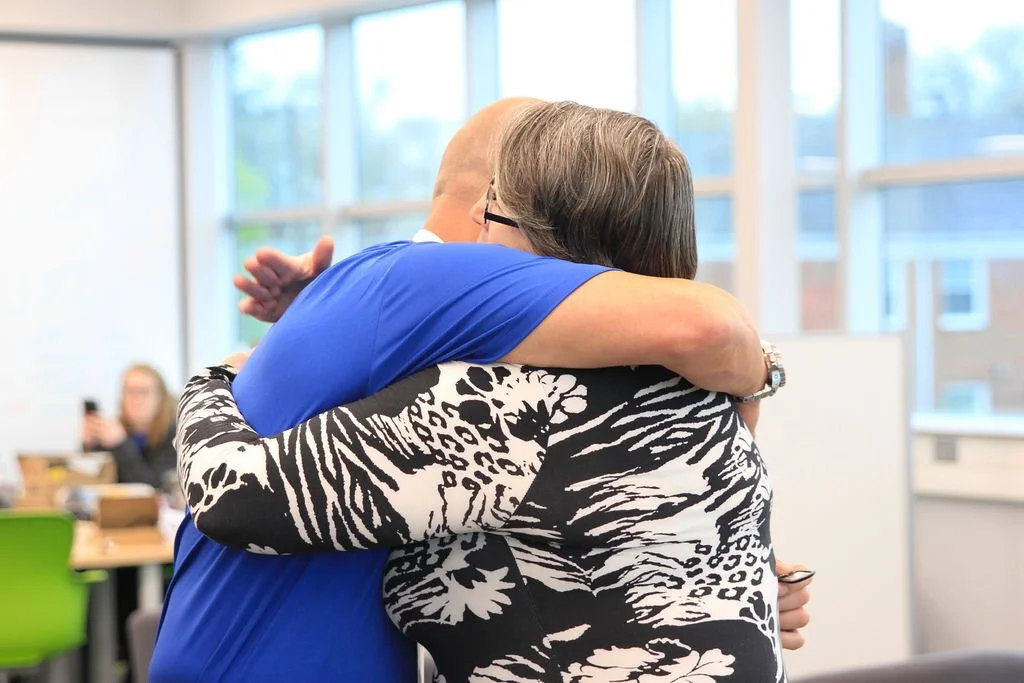 Two people hugging each other warmly in an indoor setting with large windows, one wearing a blue shirt and the other in a black and white floral patterned shirt.
