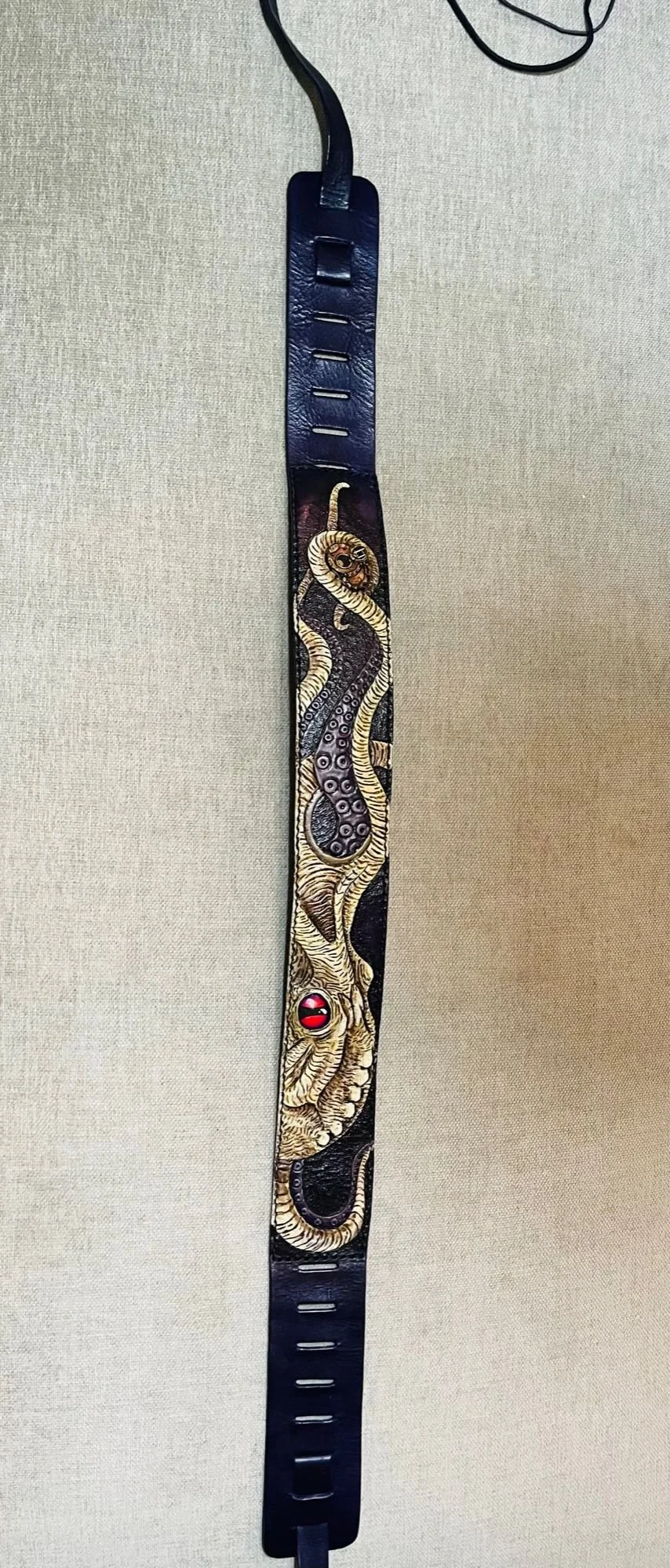 A leather belt with a dragon design, featuring a red gemstone eye and gold accents, hanging vertically against a plain background.