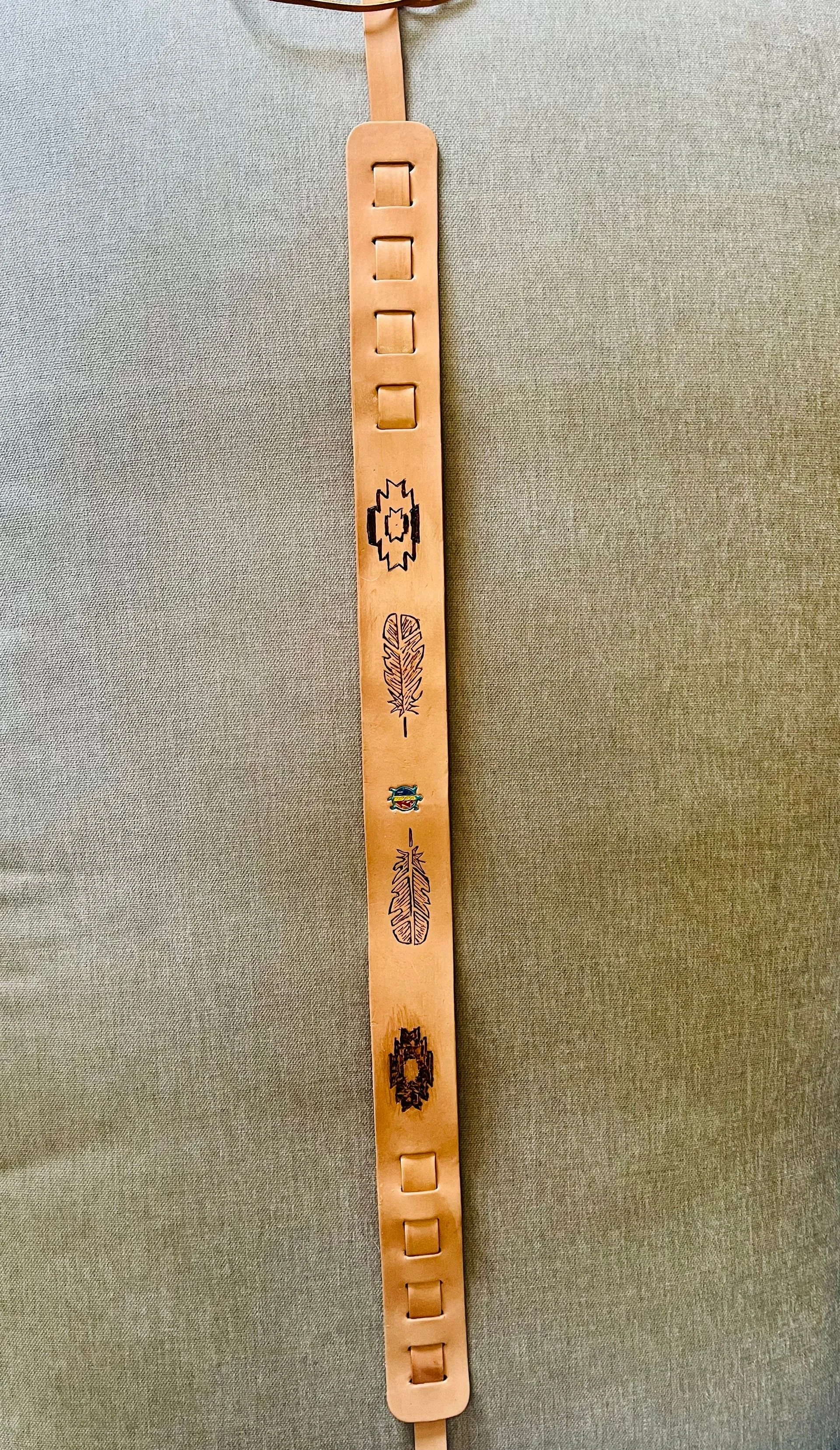 A leather strap with engraved and colored designs, including feathers and geometric patterns, hanging against a fabric background.