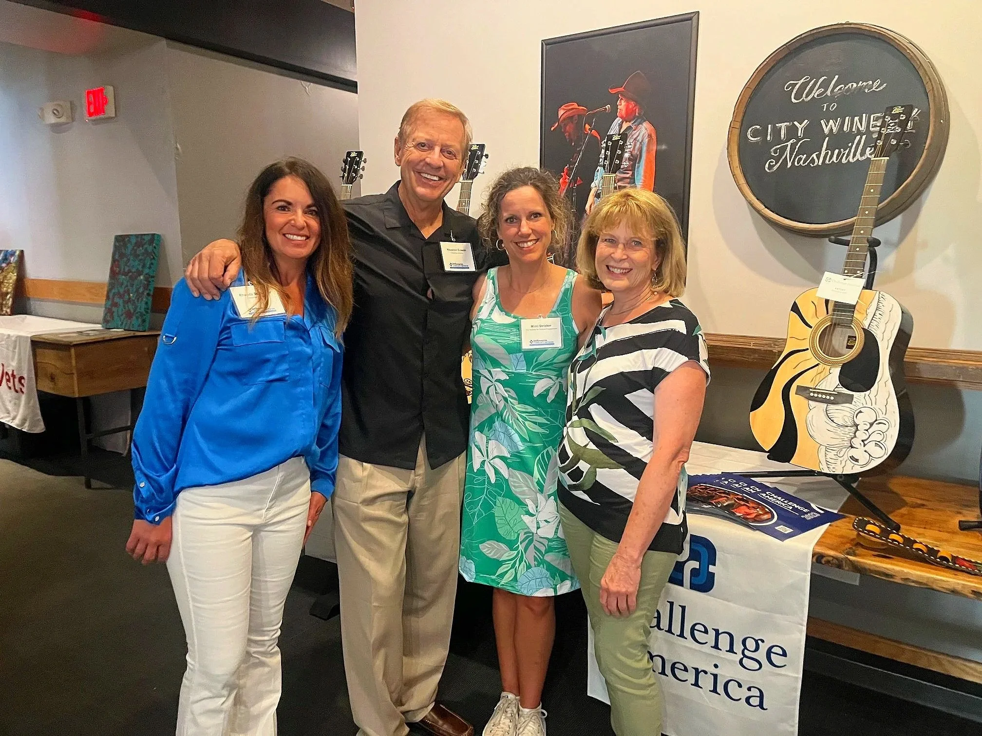 Four people standing together at an indoor event, smiling at the camera. Behind them, there is a sign that says 'Welcome to City Wine Nashville,' a guitar, and a painting. Two women and two men, all wearing casual and business casual attire.