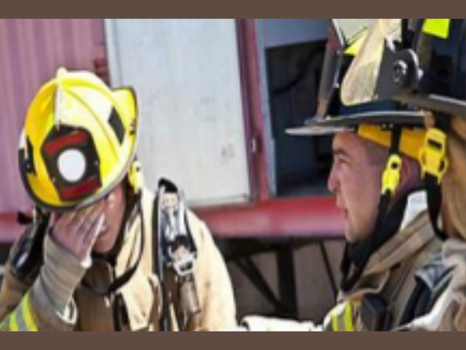 Two firefighters in gear inside a fire station, wearing yellow helmets with black and red accents, and beige firefighting uniforms, engaging in conversation.