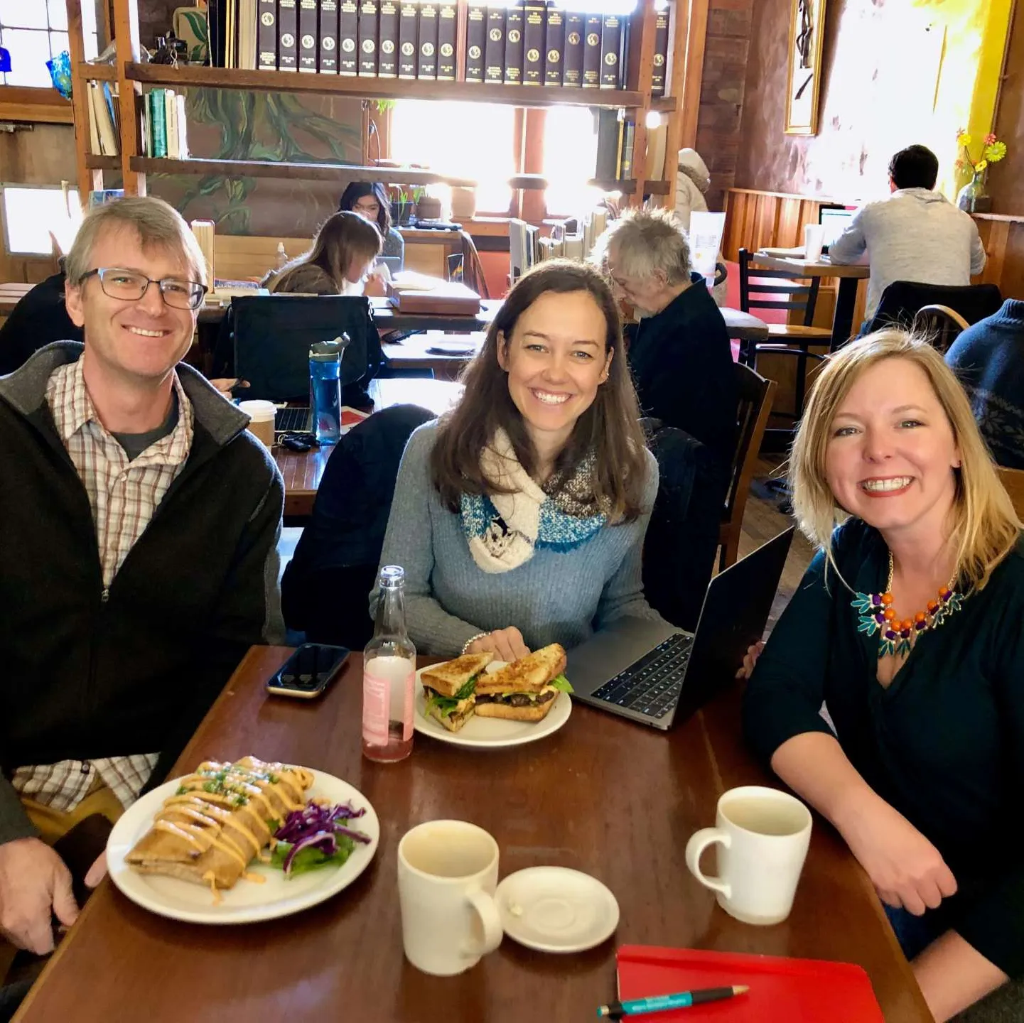 Three smiling people sitting at a table in a cozy café, with plates of food including a sandwich and a dessert, cups, and a laptop on the table, and others working in the background.