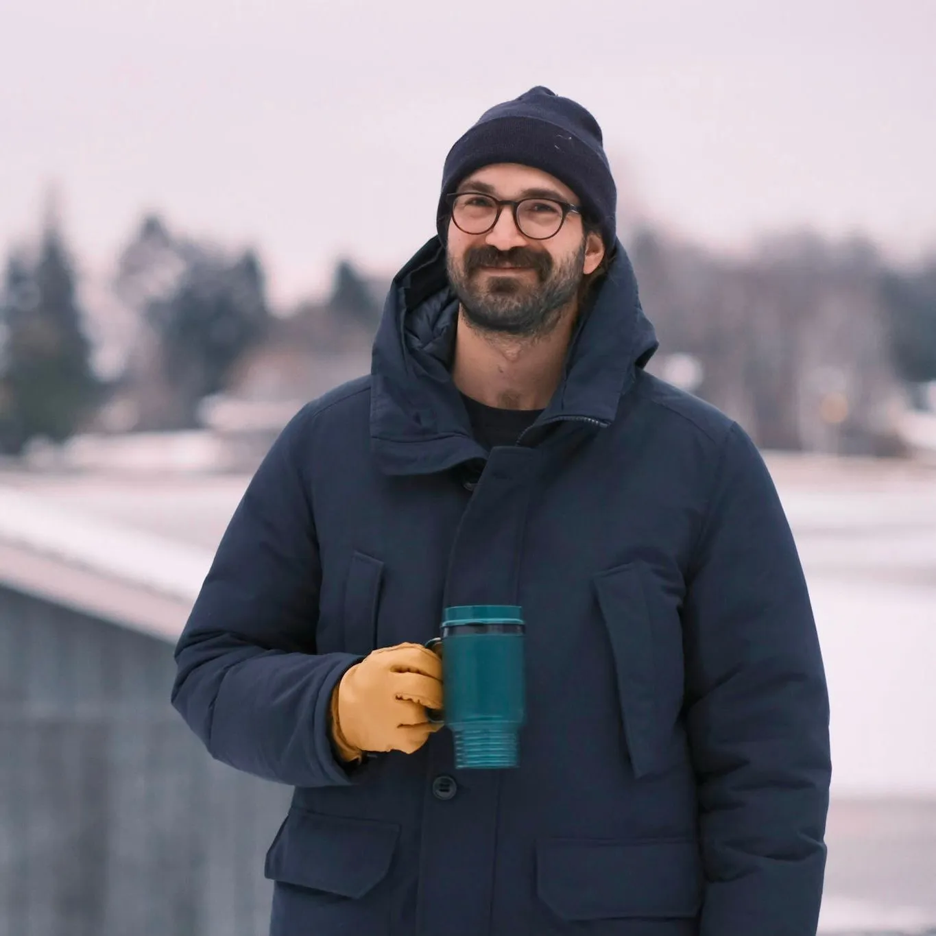A man dressed for cold weather, wearing glasses, a navy beanie, and a navy winter coat, holding a teal travel mug, standing outdoors in a snowy landscape.