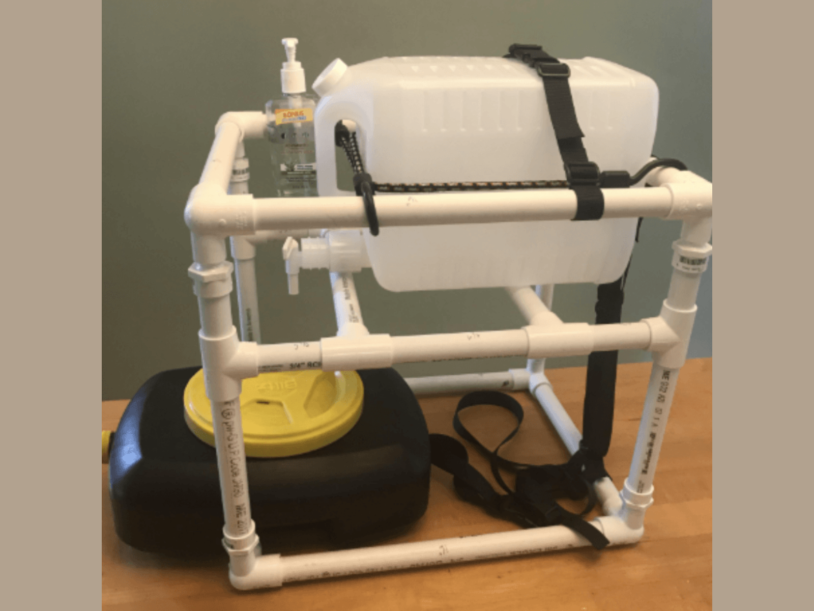 A DIY watercraft made from PVC pipes, with a white water tank, a bottle of hand sanitizer, a yellow floating device, and black straps, assembled on a wooden surface.