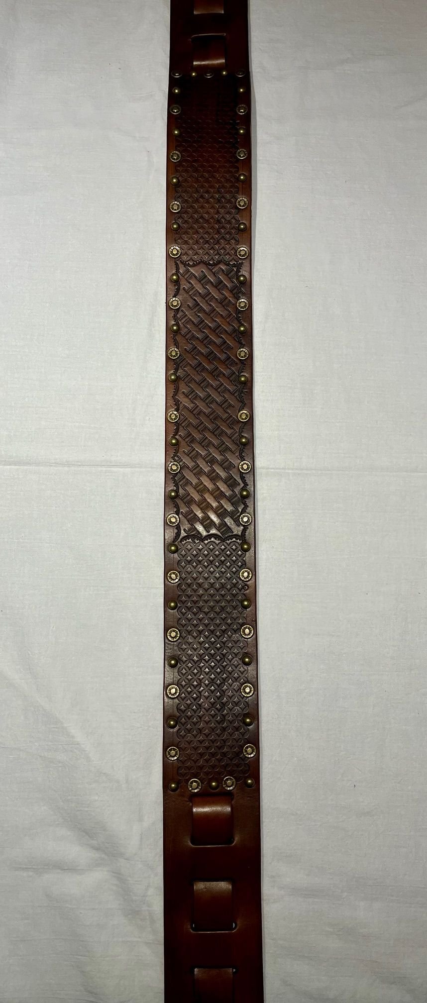 A decorative leather belt with intricate embossed patterns and metal studs, laid out flat on a light-colored surface.