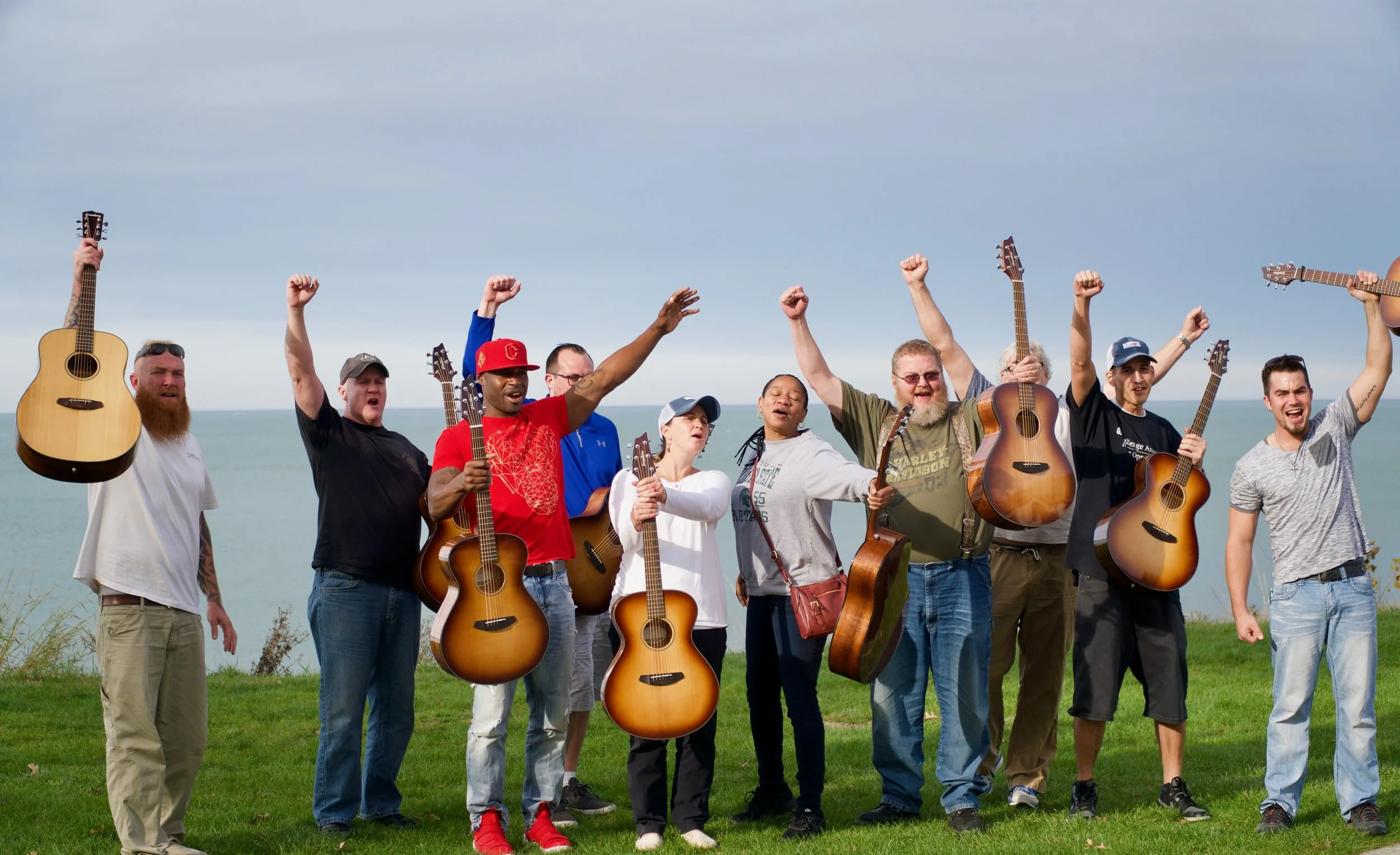 A group of people holding guitars and celebrating outdoors near a body of water, with some raising their fists in the air.