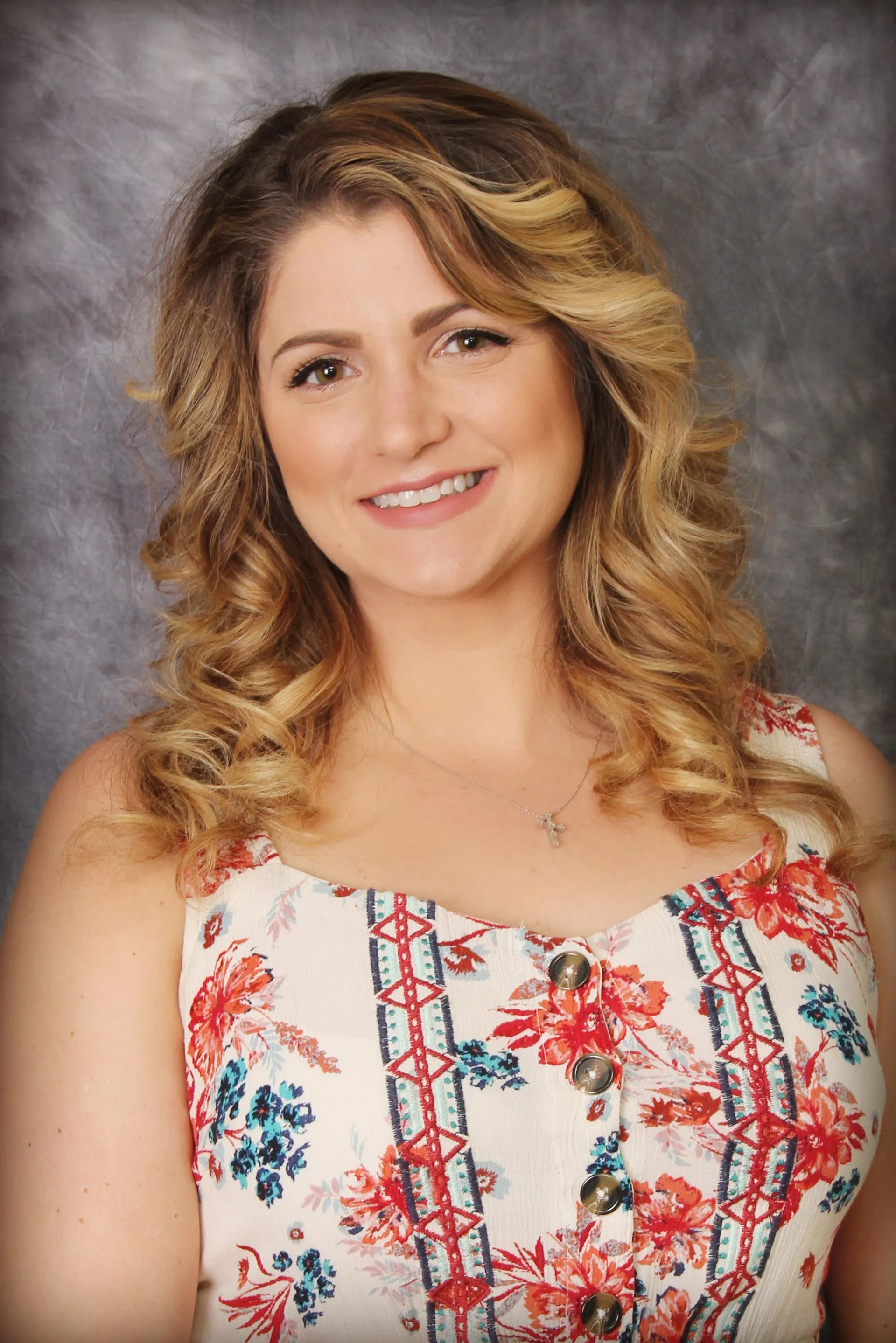A woman with wavy blonde hair, wearing a floral sleeveless top with decorative buttons and a silver necklace, smiling at the camera against a gray backdrop.
