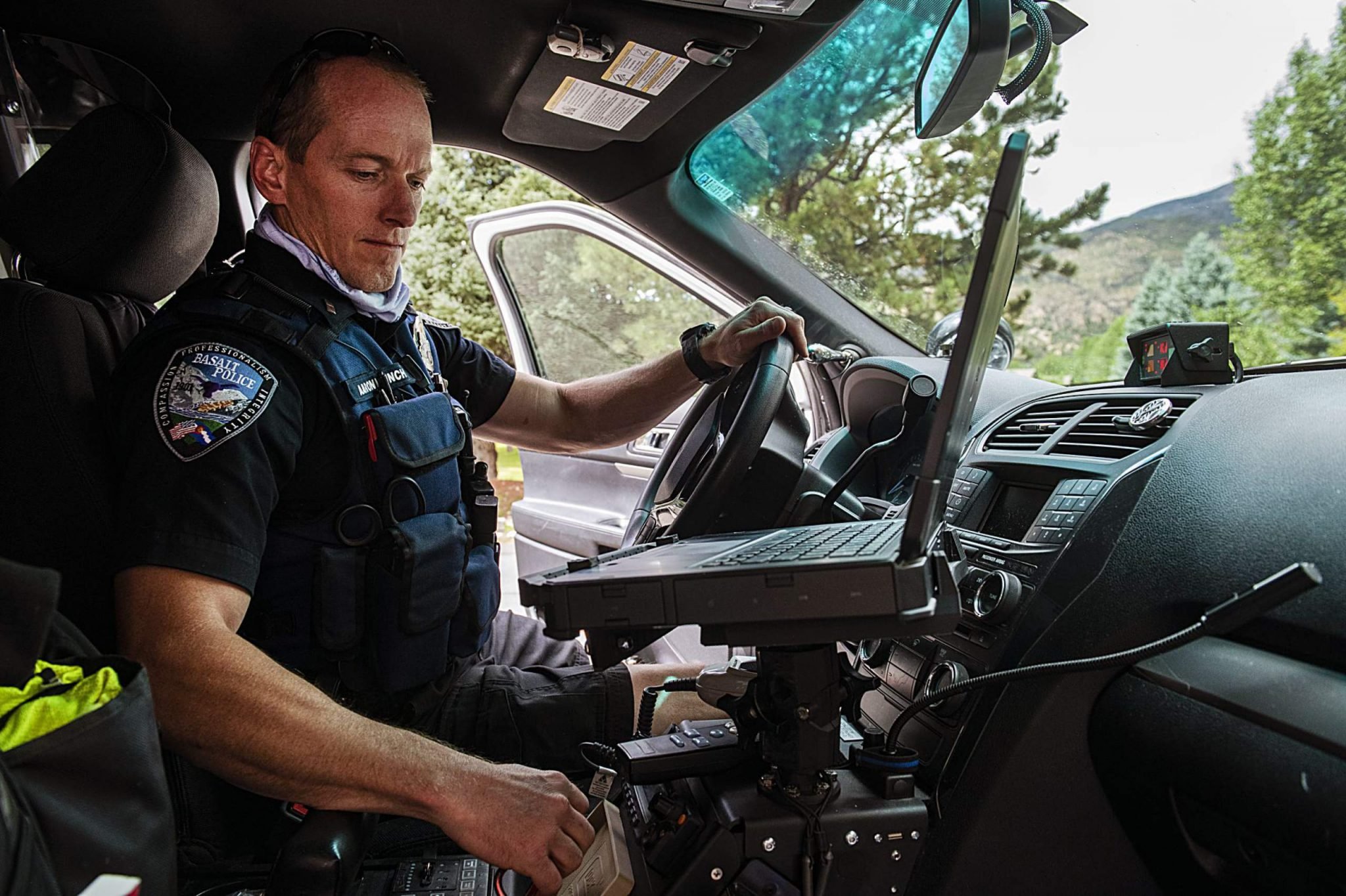 A police officer inside a patrol vehicle, looking at a laptop mounted on a stand in the front seat, with greenery and trees visible outside through the windshield.