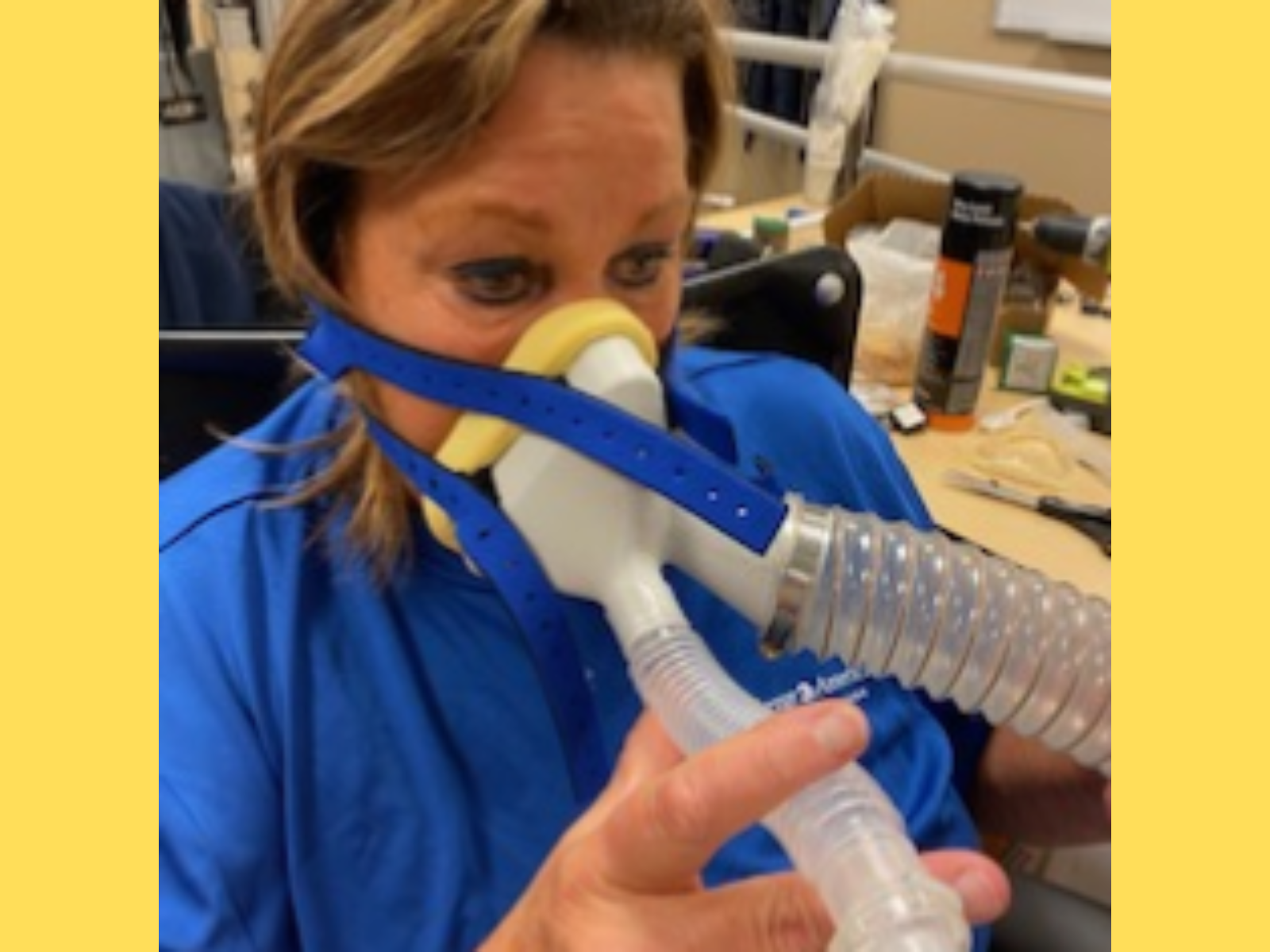 A woman wearing a blue shirt is using a respiratory mask with a transparent corrugated tube for breathing, likely inside a workshop or medical setting with tools and supplies scattered on a table in the background.