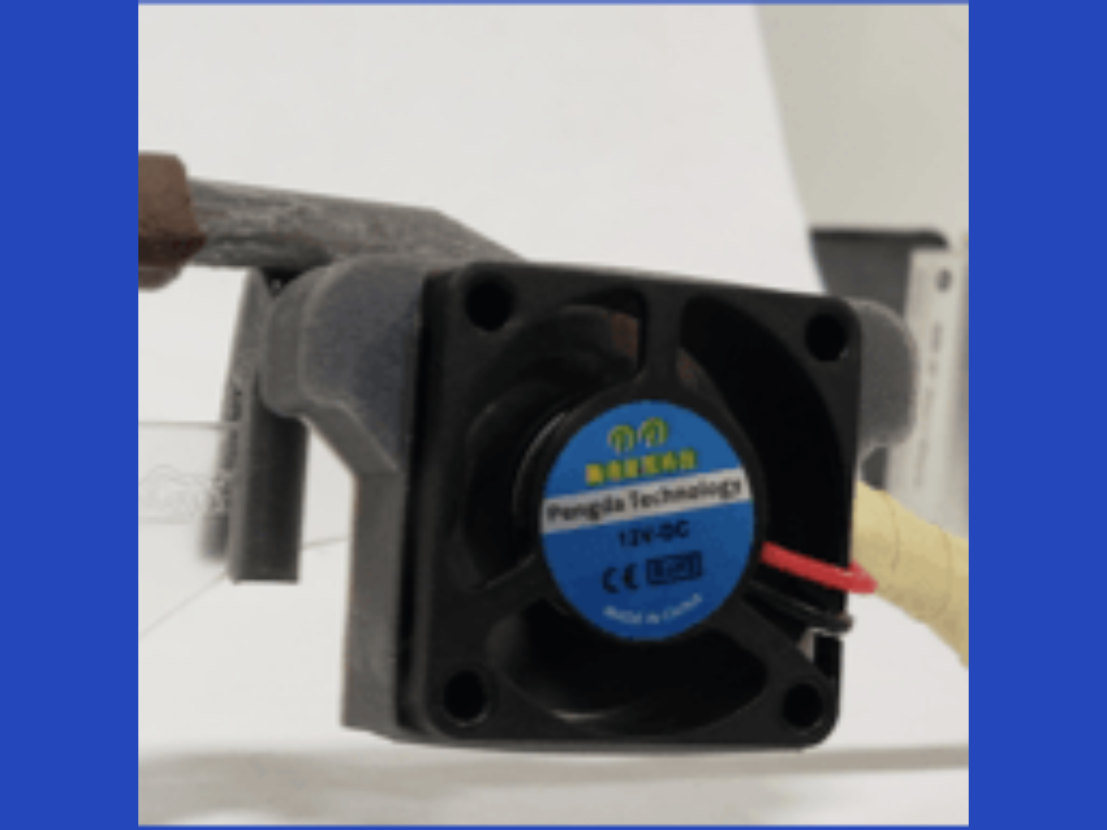 Close-up of a small black cooling fan with a blue label, mounted on a gray metal assembly, with a red wire connected to it.