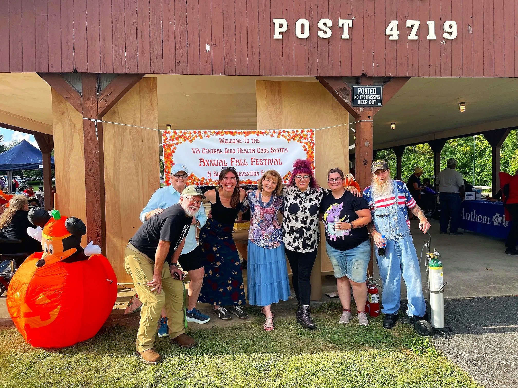Group of seven people standing in front of a sign at a fall festival, with some wearing Halloween costumes and decorations like a pumpkin with Mickey Mouse on it nearby.