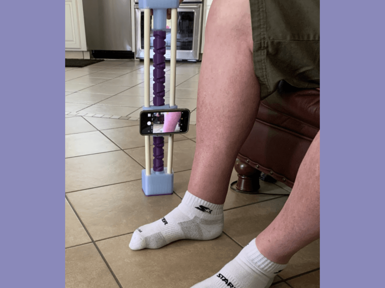 A person with a white sock on their foot is sitting on a leather chair in a kitchen. The person is holding a crutch with a purple foam grip, and a smartphone is attached to the crutch, capturing an image of their own leg.