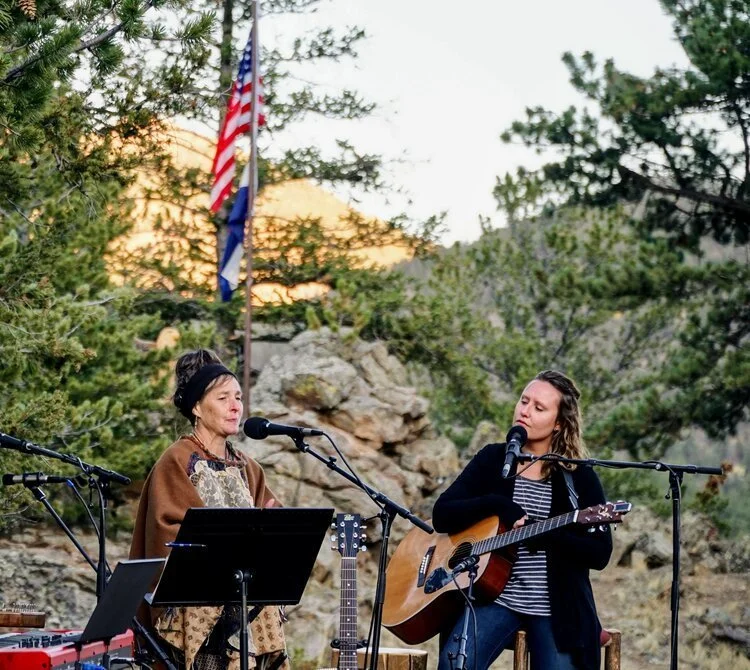 Two women perform music outdoors with microphones, one playing an acoustic guitar and the other singing, surrounded by trees and rocks, with American flags in the background.