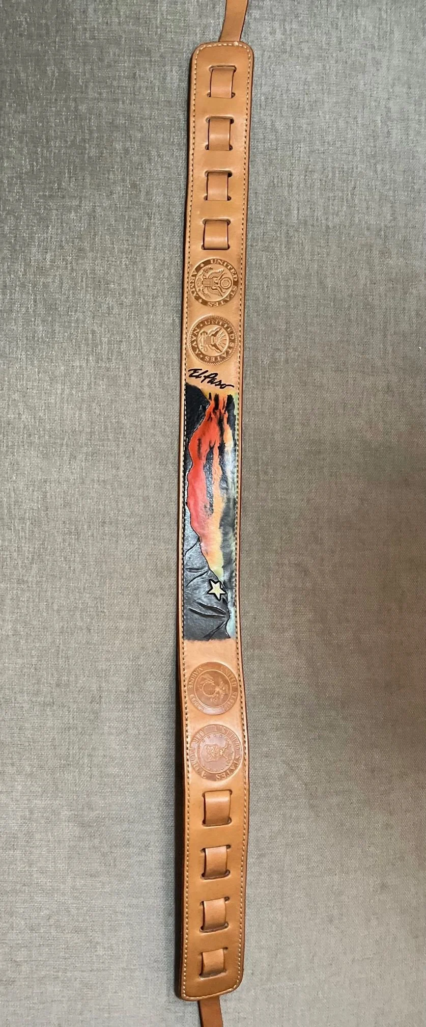 Leather belt featuring several embossed badges and a painted landscape with mountains, sunset, and trees, with the word 'El Paso' written on it.