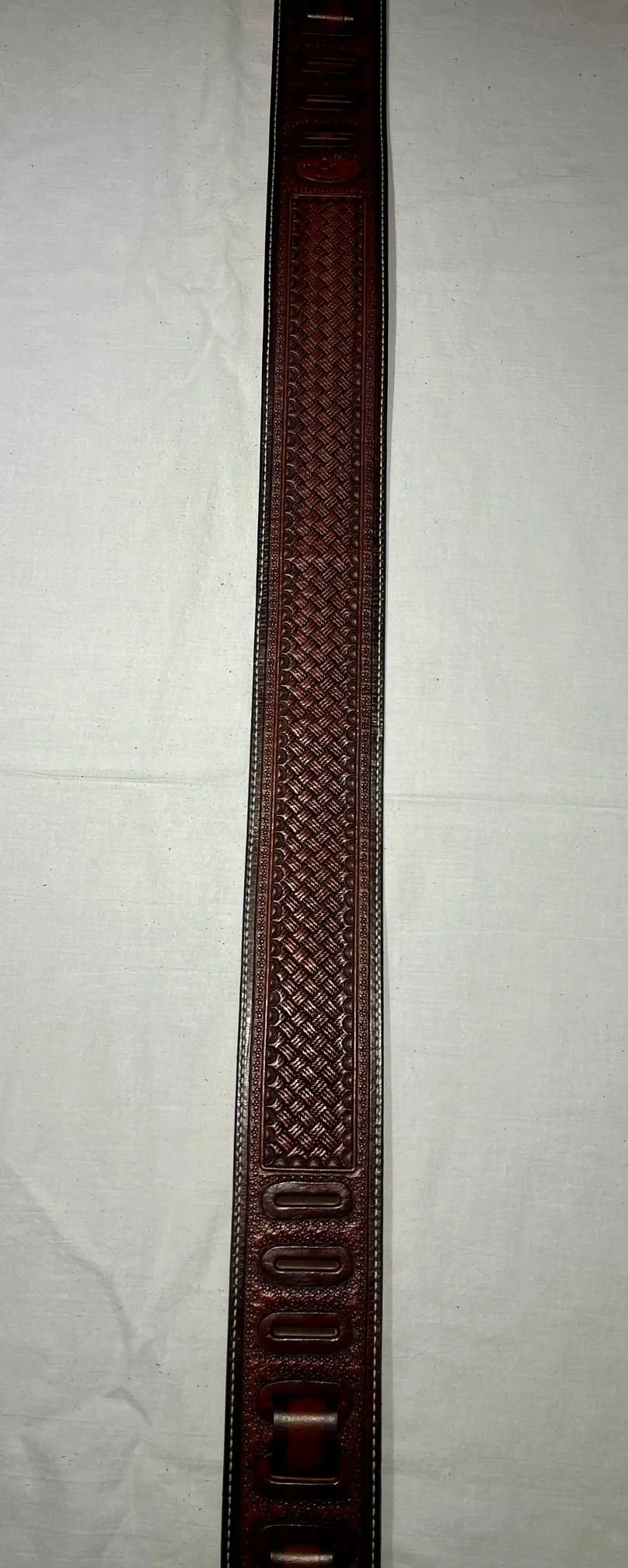 A long, narrow, leather watch strap with crosshatch and stamped patterns, and multiple adjustment holes, laid out on a light-colored fabric background.