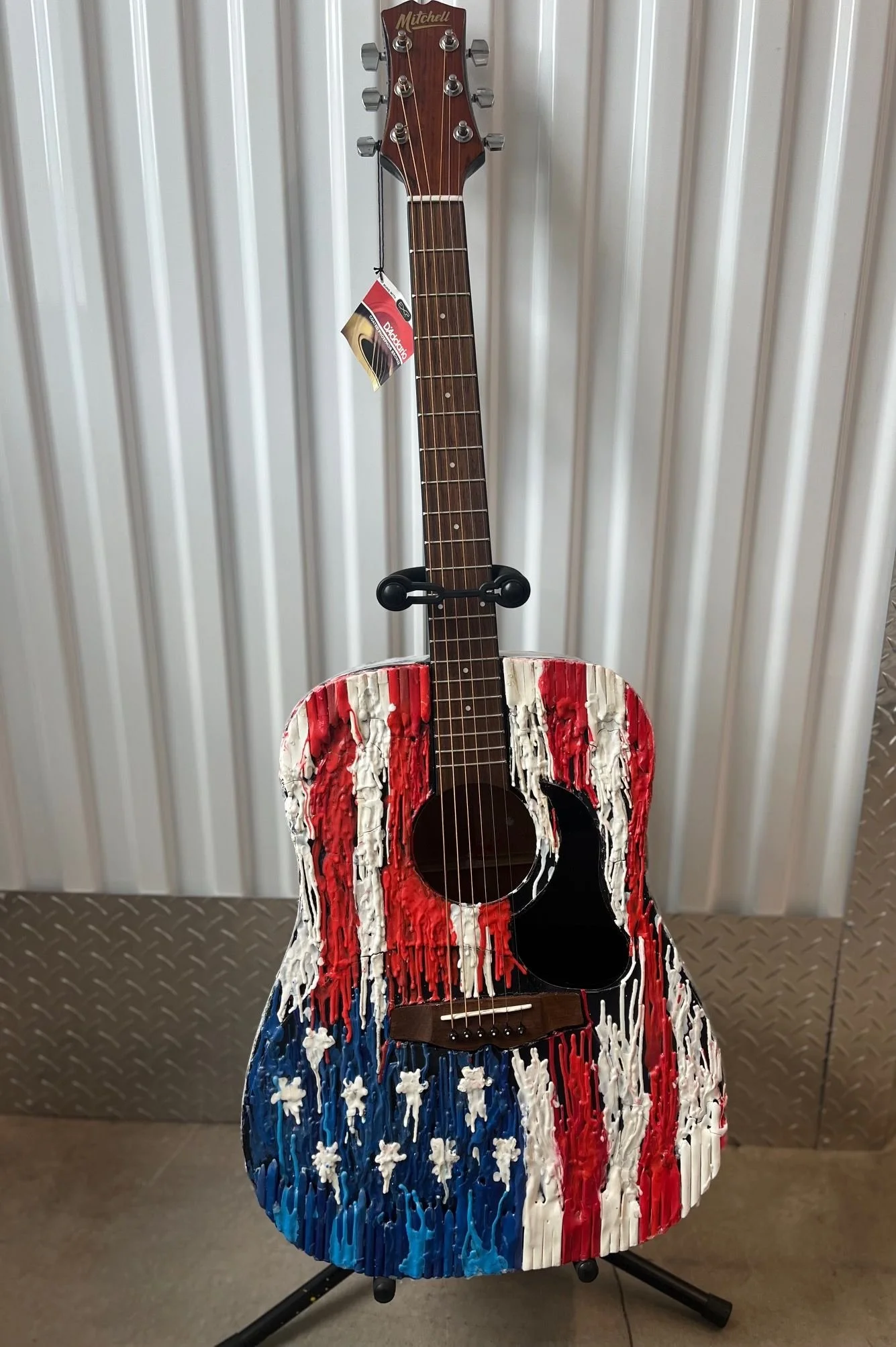 An acoustic guitar with a body painted with an American flag design, featuring red, white, and blue colors with white stars. The guitar is on a stand against a white wall with vertical panels.