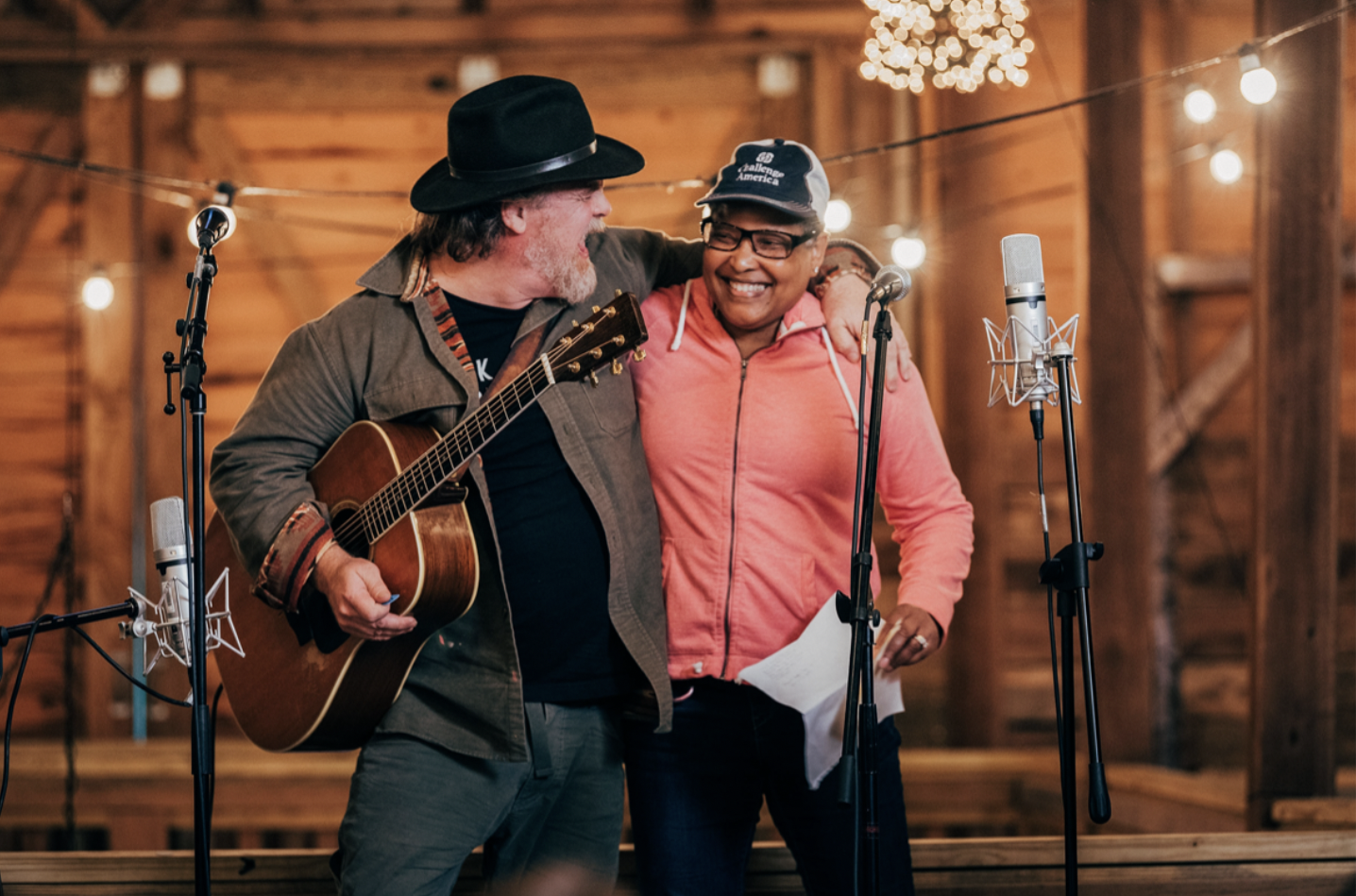 Two people are singing and playing guitar on stage in a rustic wooden barn. They are smiling and have their arms around each other, surrounded by microphones and warm string lights.