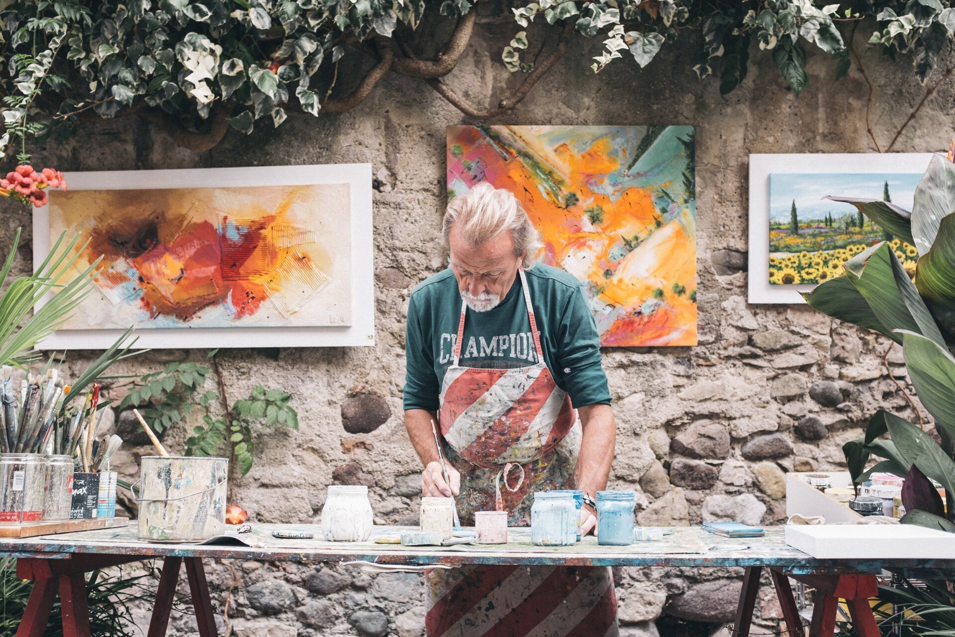 An elderly man painting in his art studio, with abstract paintings on the stone wall behind him and plants on either side, a table with paint jars and brushes in front.