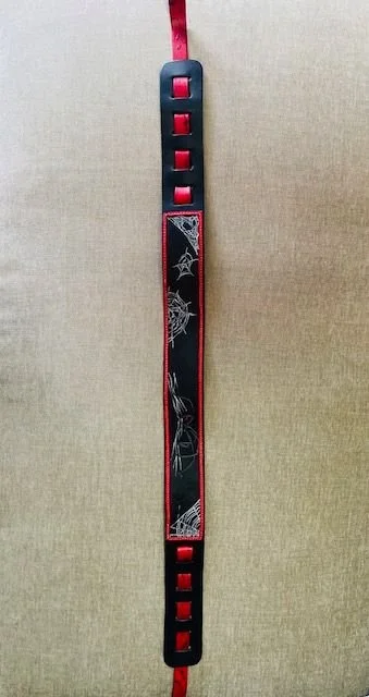 A black and red bracelet with a spider web and spider design on it, hanging vertically against a beige background.