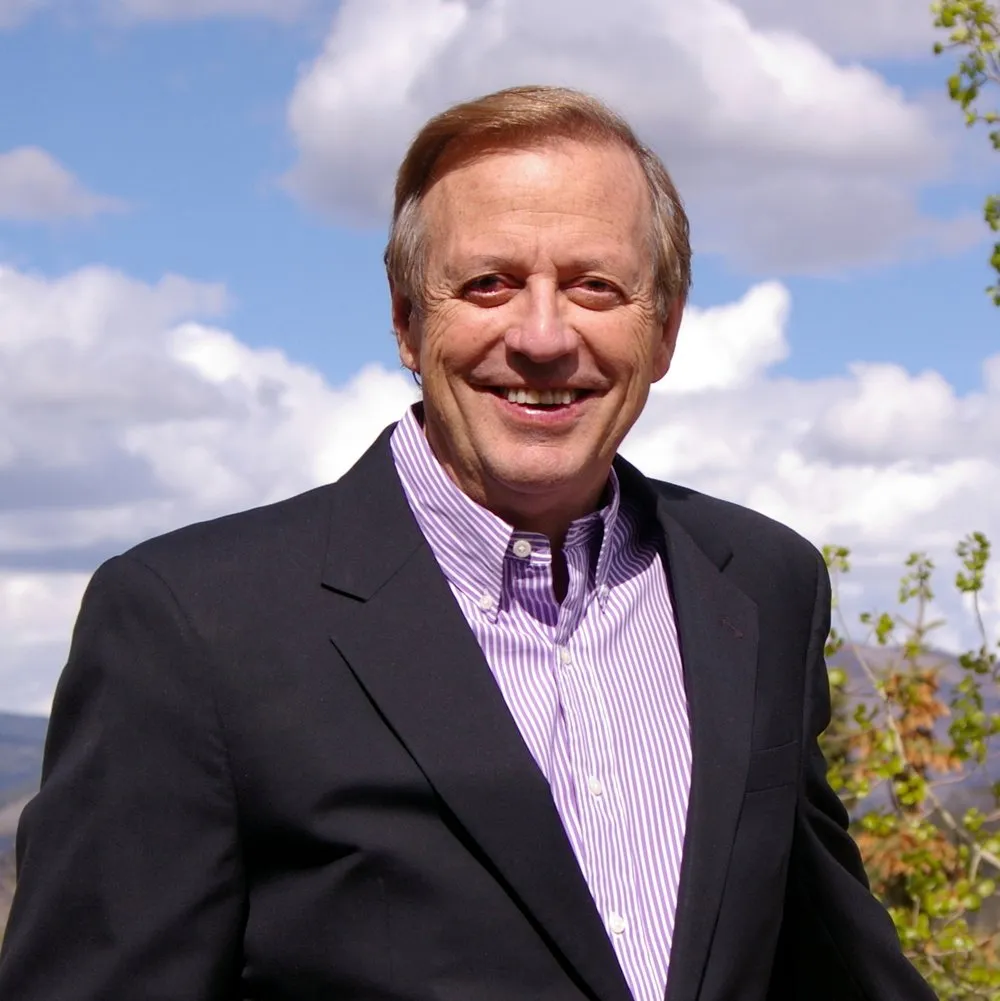 A smiling man in a black blazer and purple striped shirt outdoors with a cloudy sky and greenery in the background.