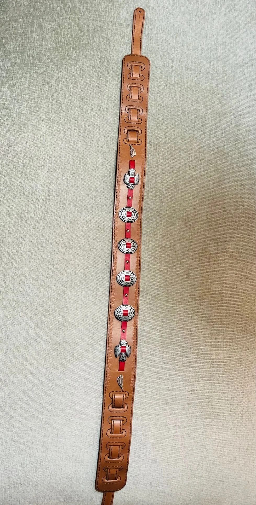 A tan leather belt with decorative silver and red embellishments, including small shields and a central emblem, on a light fabric background.