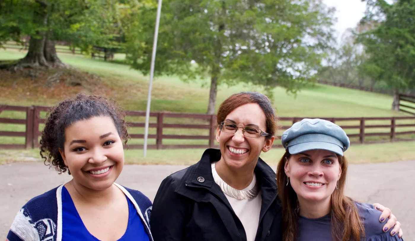 Three women smiling outdoors, standing on a road with a wooden fence and trees in the background.