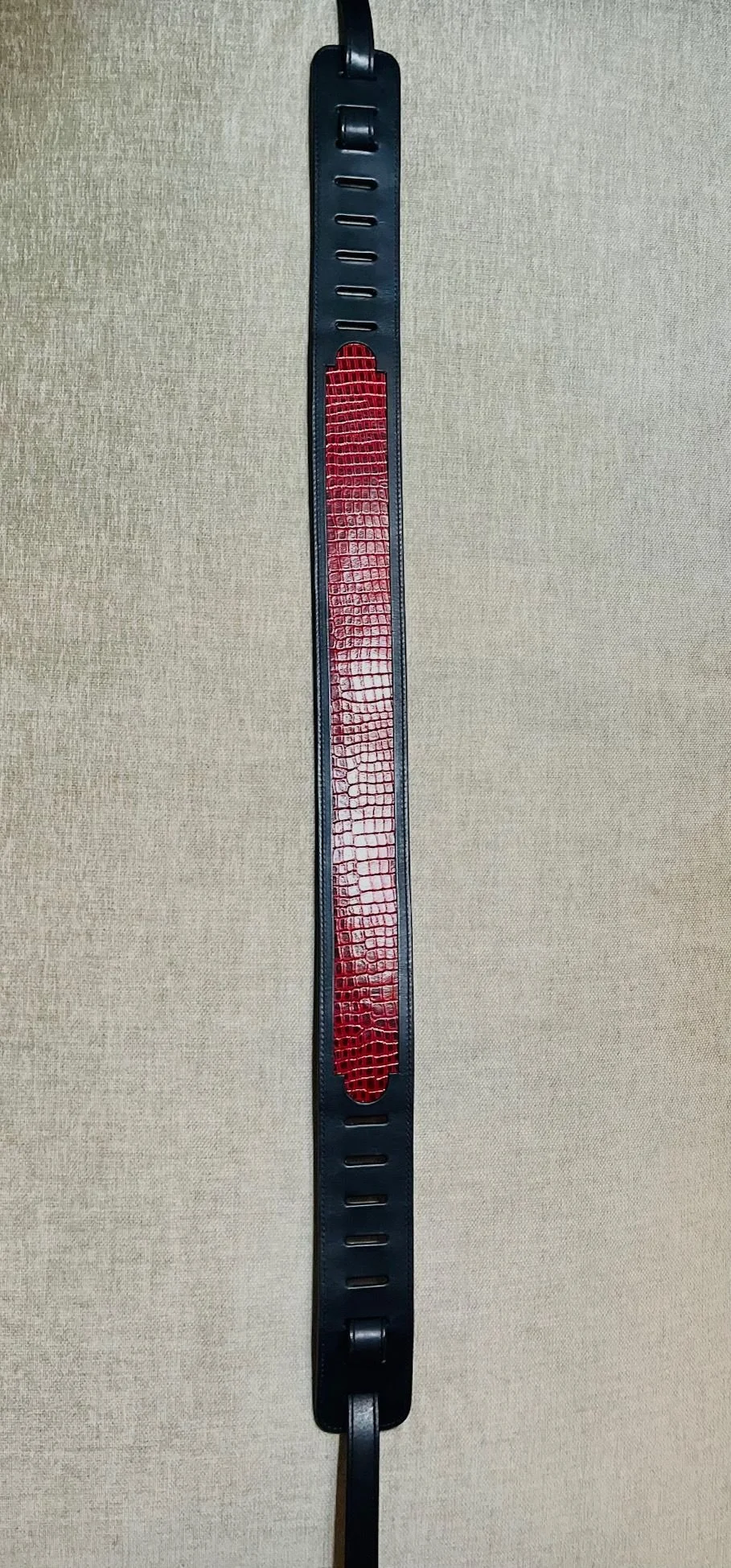 A black leather belt with a red and black crocodile pattern in the center, laid flat against a beige textured background.
