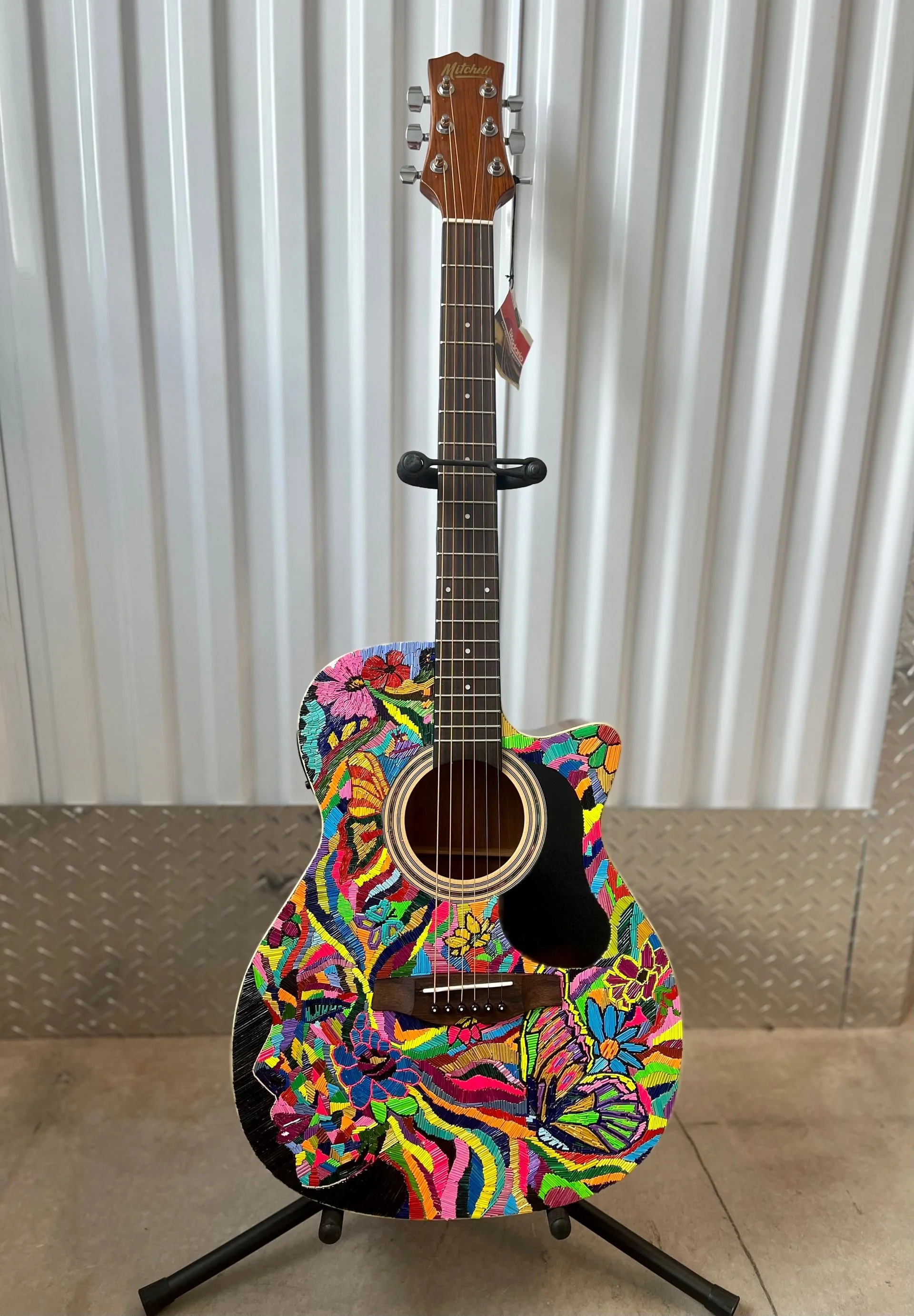 Colorful, floral-patterned acoustic guitar on display, standing on a black guitar stand against a wall.