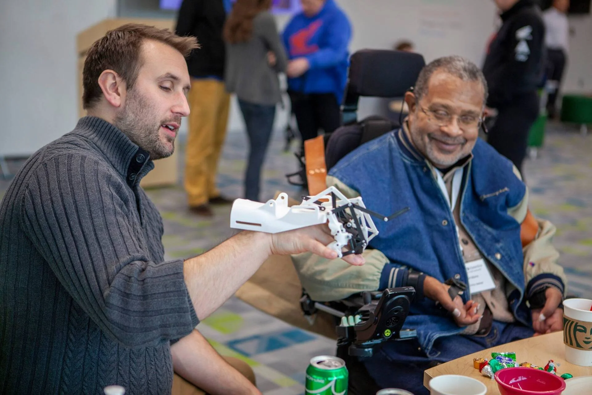 A young man demonstrating a robotic device to an older man with a smile at a table with snacks and drinks in a busy indoor setting.