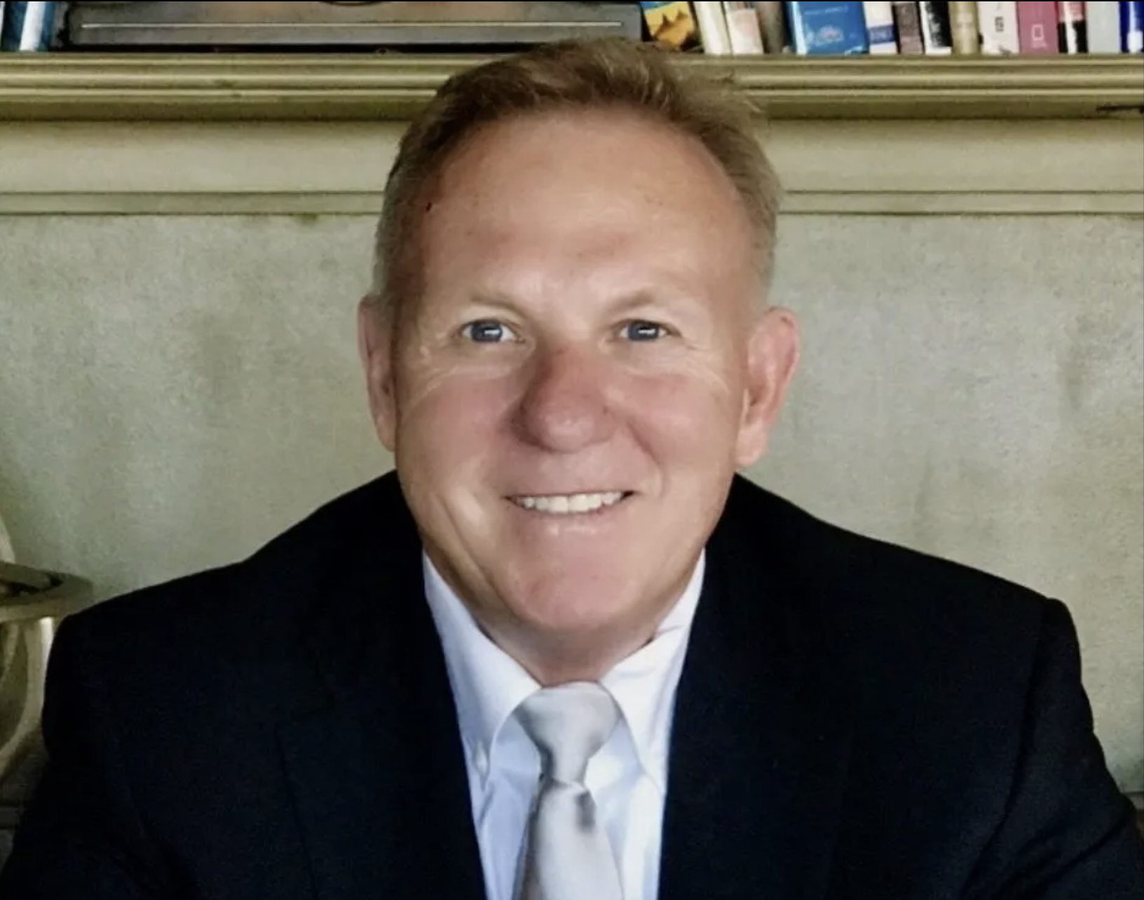 A man with light skin, short red hair, and blue eyes wearing a black suit and a light-colored tie, smiling at the camera. He is in front of a neutral wall with a bookshelf partially visible in the background.