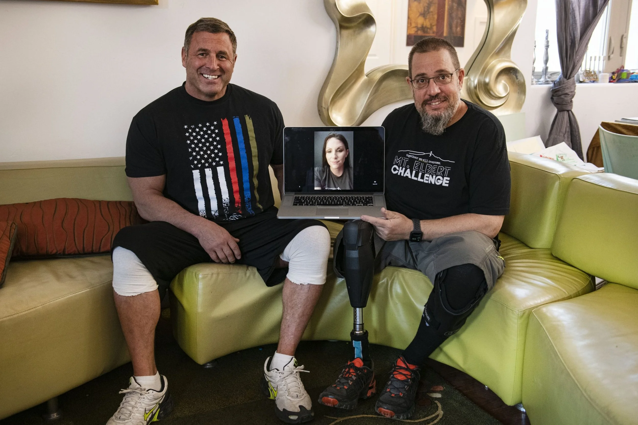 Two men sitting on a yellow couch, smiling at the camera, with a woman on a video call displayed on a laptop. They are in a living room with decorative wall art, curtains, and miscellaneous items on a side table.