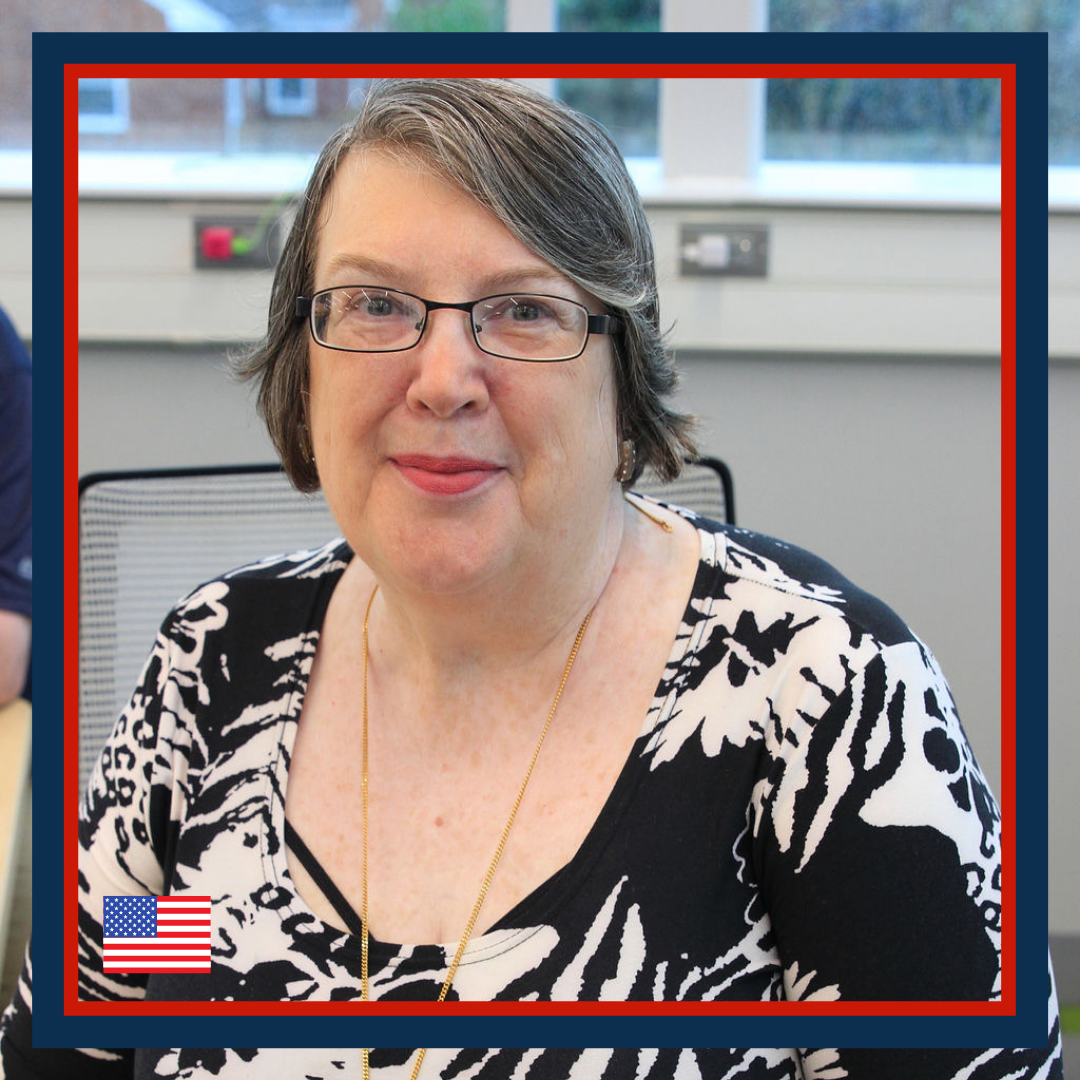 A middle-aged woman with short gray hair, glasses, and wearing a black and white patterned top, sitting indoors near a window.