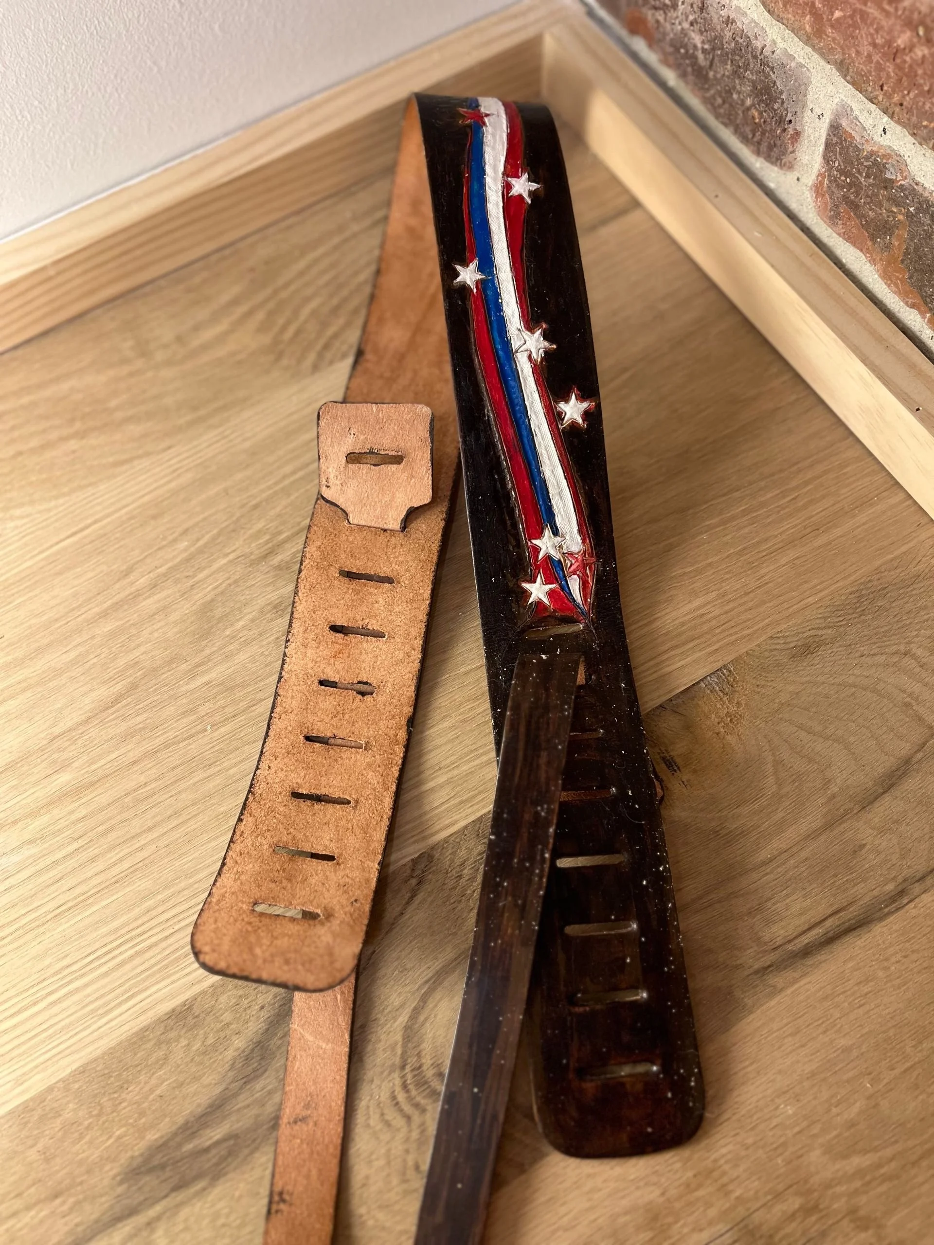 A black wooden slapstick with a patriotic red, white, and blue stripe and white stars, placed on a wooden surface next to a light brown leather strap with holes.