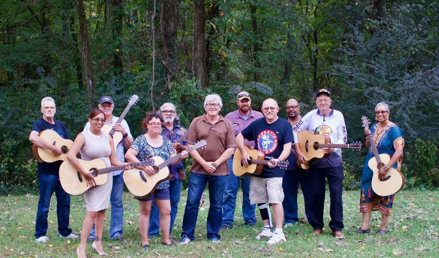 Group of eleven people standing outdoors on grass with trees in the background, some holding guitars, dressed casually.