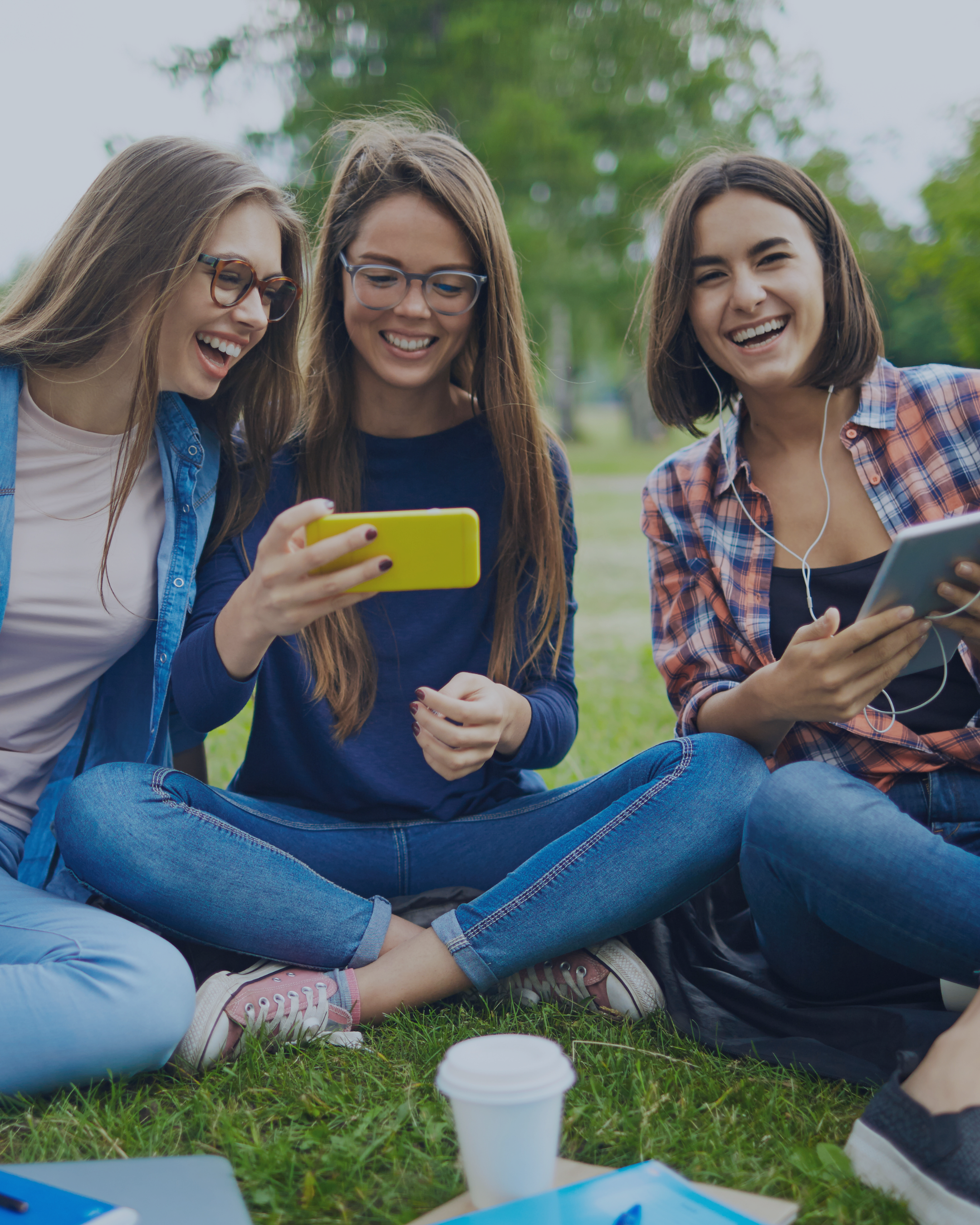 Three young women sitting on grass in a park, looking at a phone and tablet, smiling and enjoying time together.