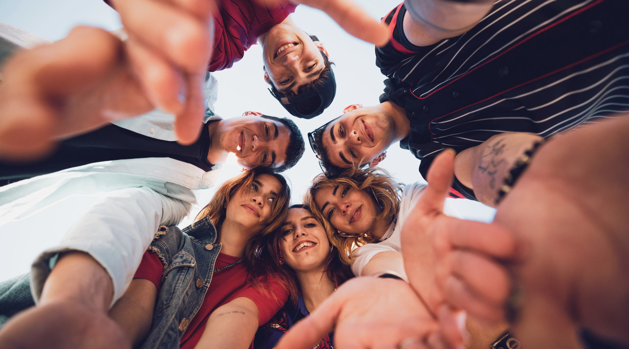 Group of seven friends looking down into the camera, smiling, as they form a circle and reach towards the camera with their hands, outdoors during the daytime.