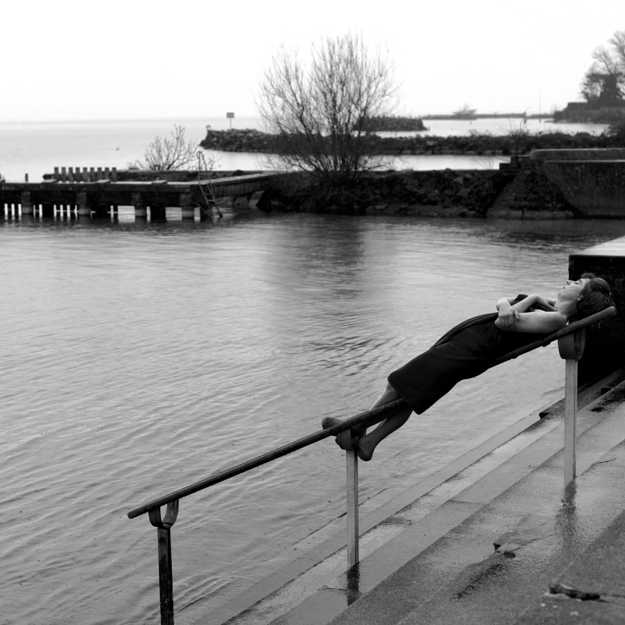 A woman reclining on a slanted railing by a body of water, with a calm expression, in black and white.