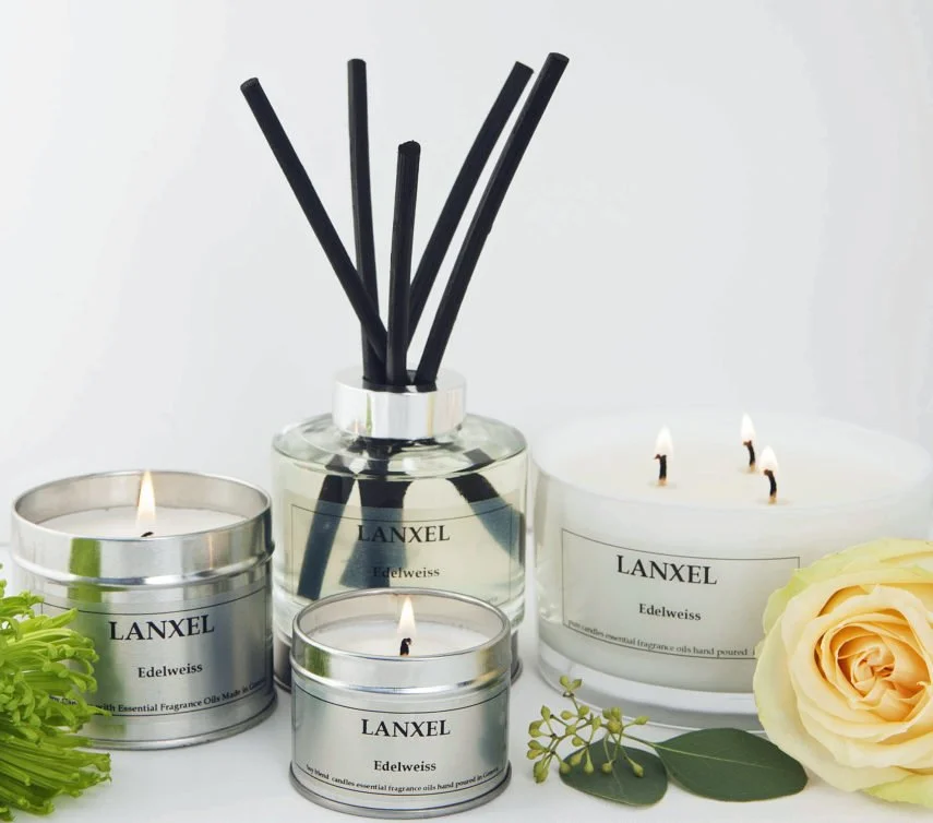 Three candles and a diffuser with black reed sticks, all labeled 'LANXEL Edelweiss,' along with a white rose, greenery, and a white background.
