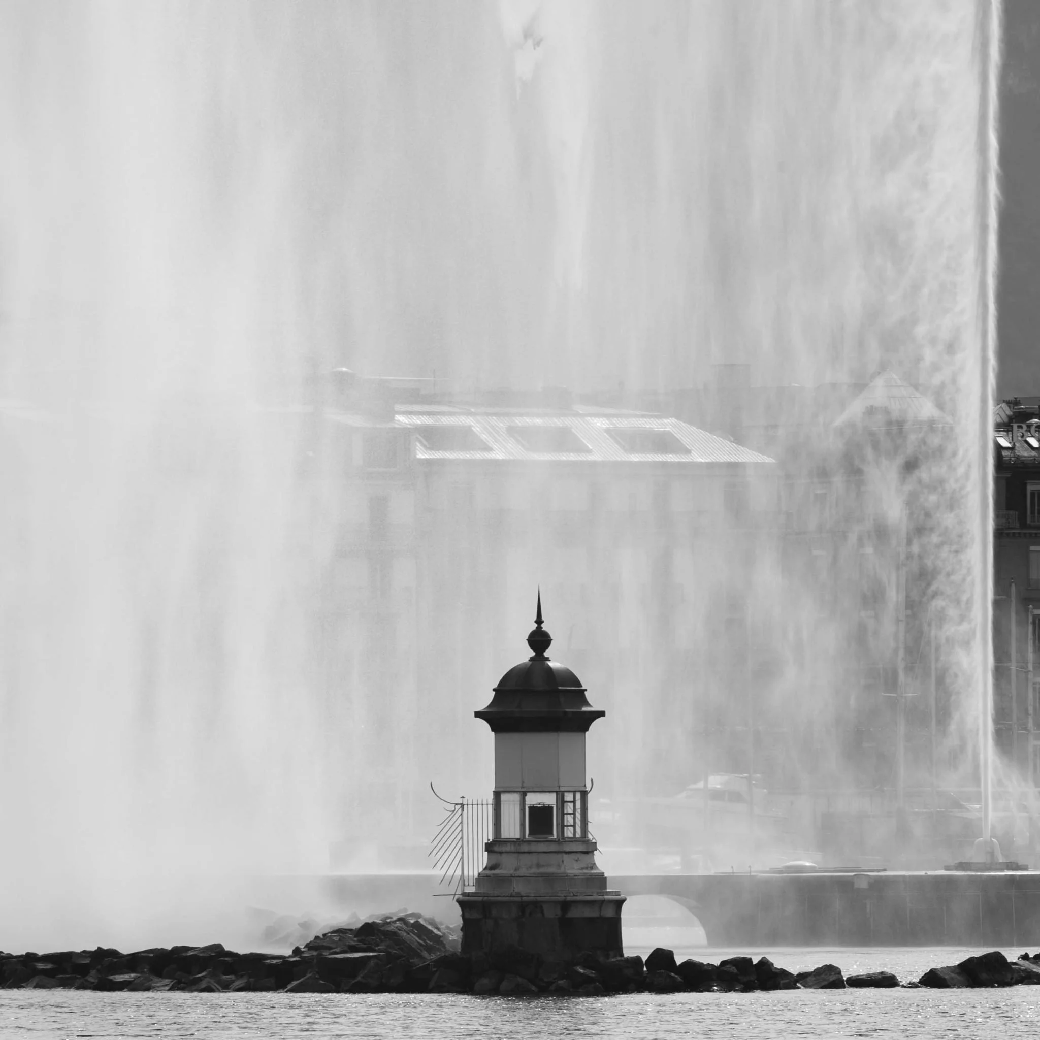 Black and white photo of a small lighthouse on a rocky outcrop in water, with a large waterfall behind it creating a misty backdrop.