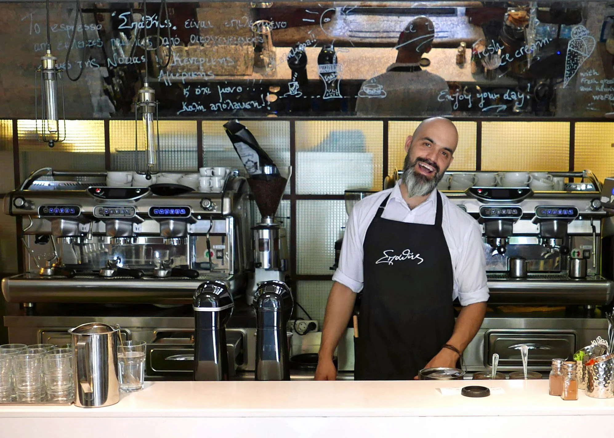 A smiling barista with a beard wearing a black apron that has white writing, standing behind a coffee counter in a café with three silver espresso machines, some jars and glasses, with a chalkboard menu and a mirror reflecting the café interior behind him.