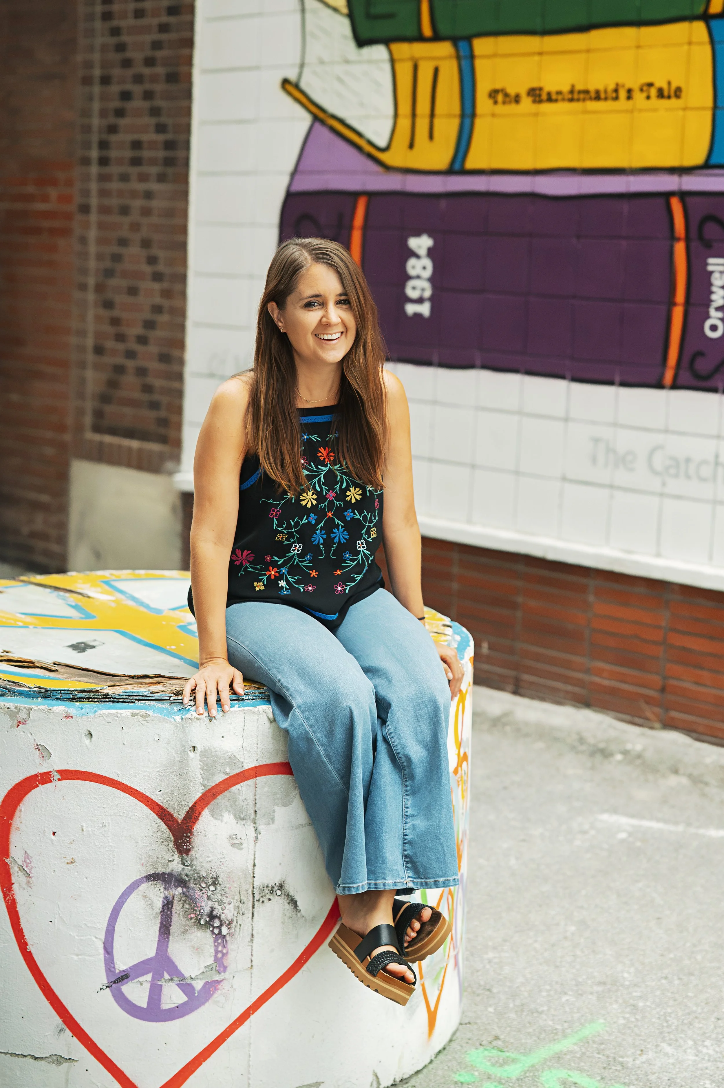 Kaitlyn sits smiling on a concrete barrier covered in colorful symbols like a heart and a peace sign. Behind her is a mural of book spines.