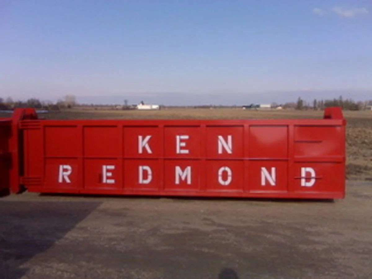 Red farm trailer with white letters spelling 'KEN REDMOND' lying on its side, set against a rural landscape with open fields and distant buildings.