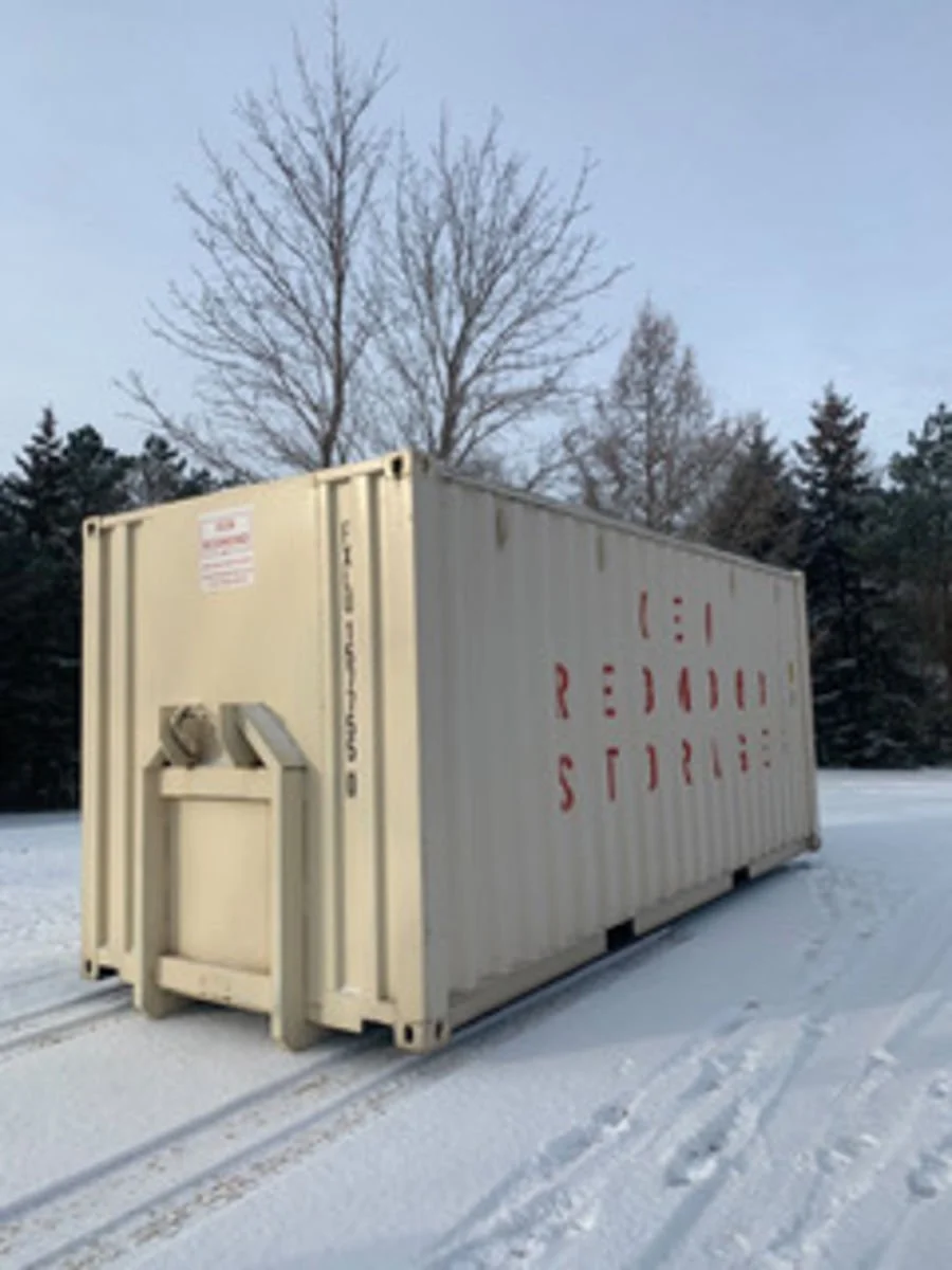A white shipping container with red lettering that reads 'REDDING STORAGE' on snow-covered ground, with bare trees and evergreens in the background under a light blue sky.