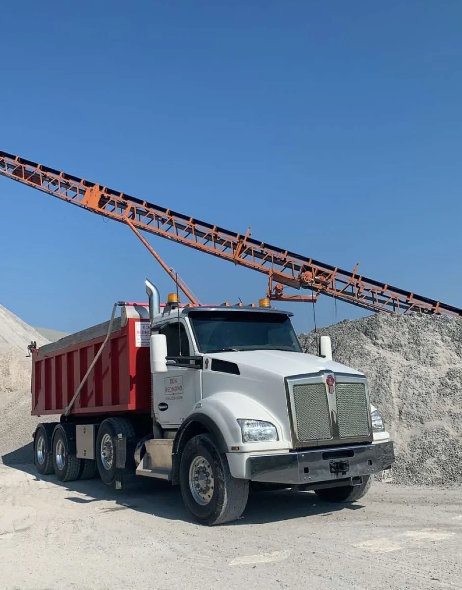 A white dump truck with a red bed parked on a gravel surface at a construction site, with dirt piles and a large orange conveyor or conveyor belt moving gravel or dirt against a clear blue sky.