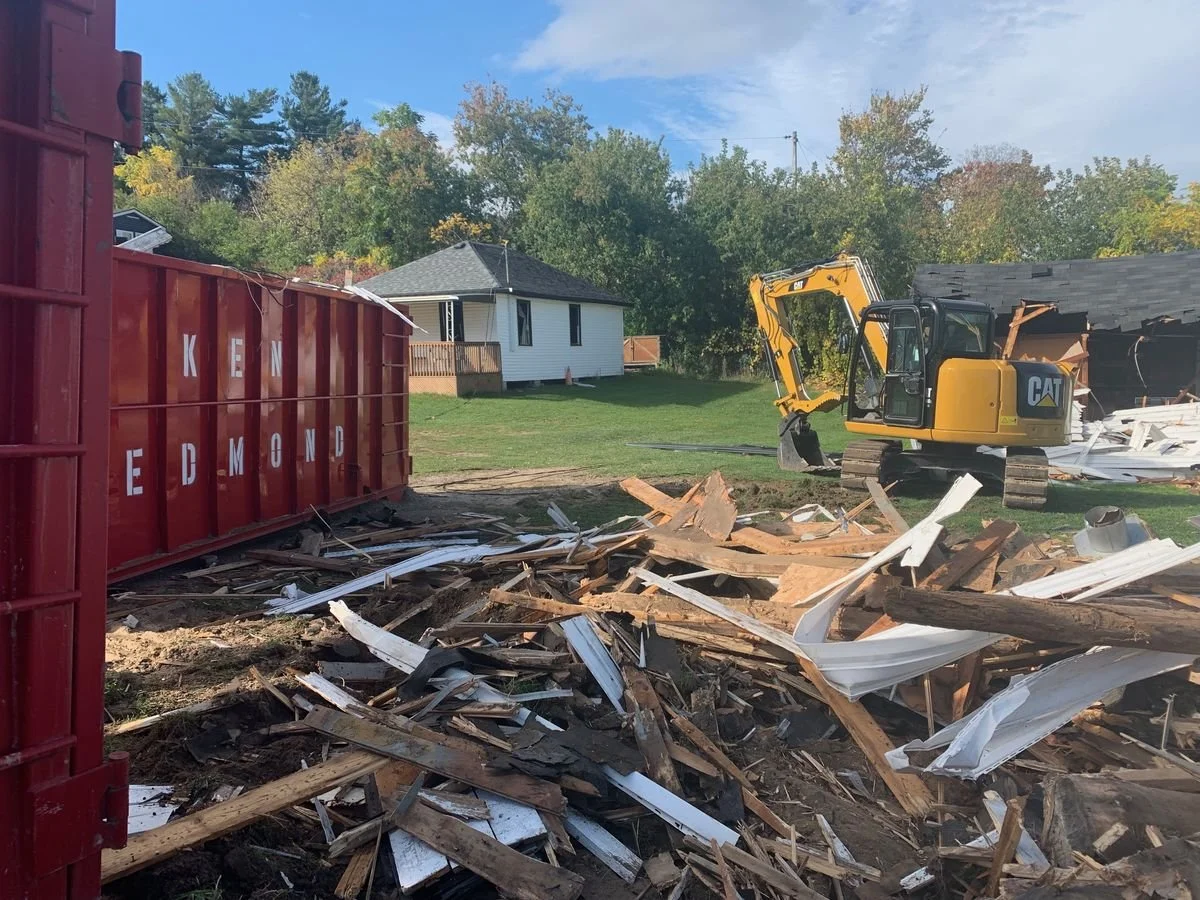 A construction site with debris, a yellow excavator, and a partially destroyed building, with a house and trees in the background.