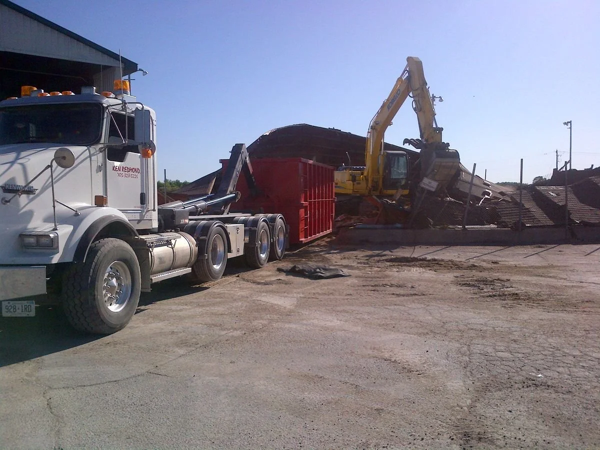 Construction site with a white semi-truck and a yellow excavator moving debris and dirt.