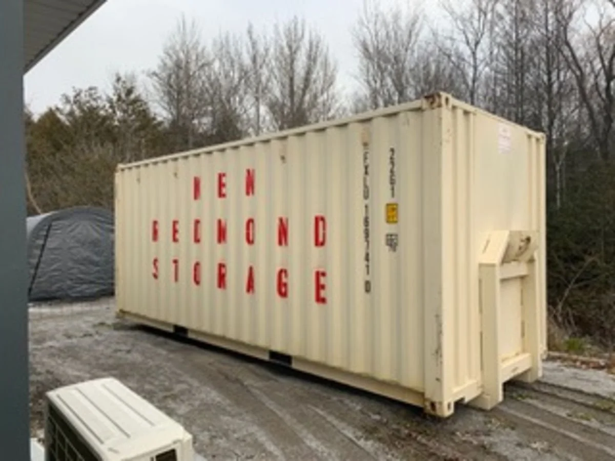 A beige shipping container with red letters spelling out "RENT SECOND STORAGE" outside on a gravel surface with trees in the background.