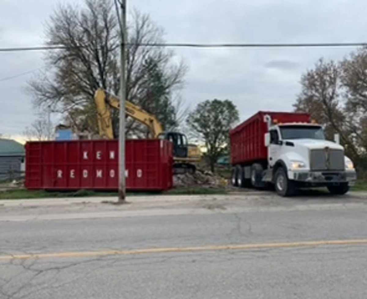 Construction scene with a yellow excavator inside a red dumpster, a white dump truck parked nearby, and trees in the background.